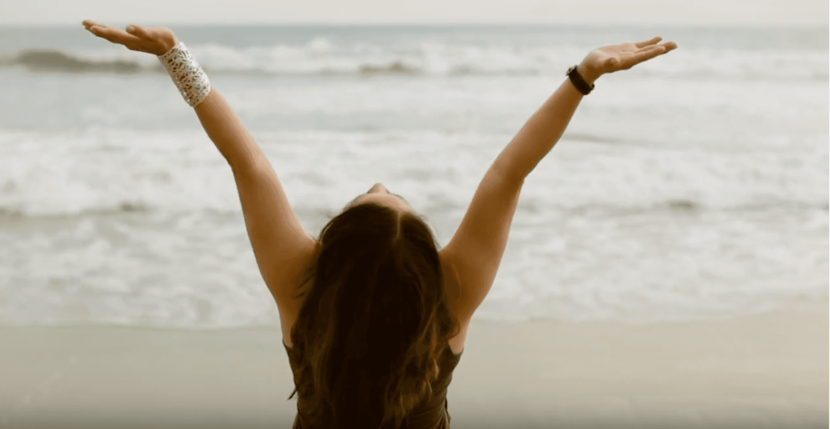 A person with long hair stands on a sandy beach facing the sea, with arms raised towards the sky in a gesture of joy or relaxation. Waves gently lap the shore in the background.