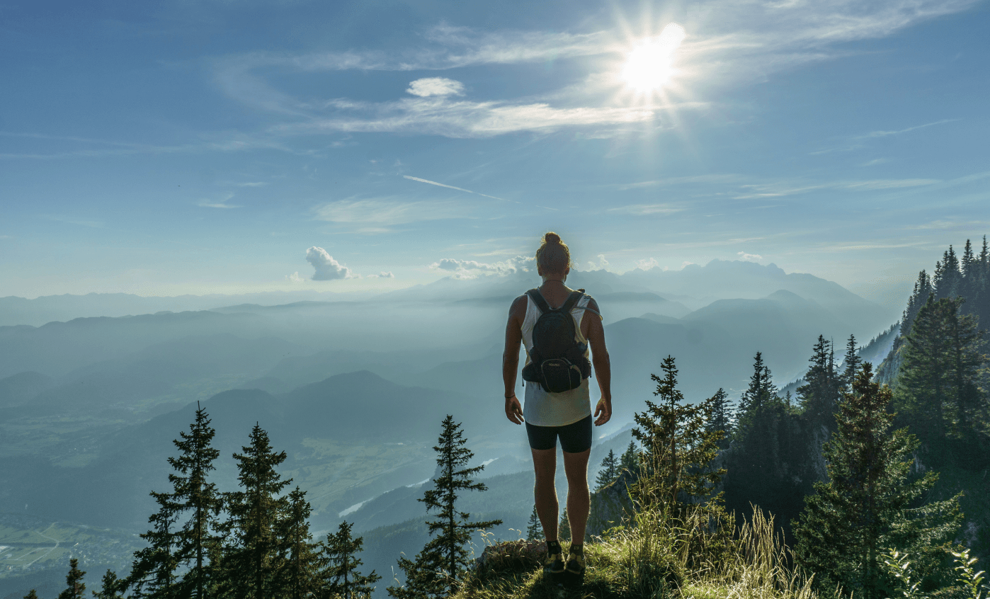 A person stands on a grassy mountain peak, wearing a backpack, gazing at a misty valley below. The sun shines brightly in a clear blue sky, casting light over distant mountains and evergreen trees.