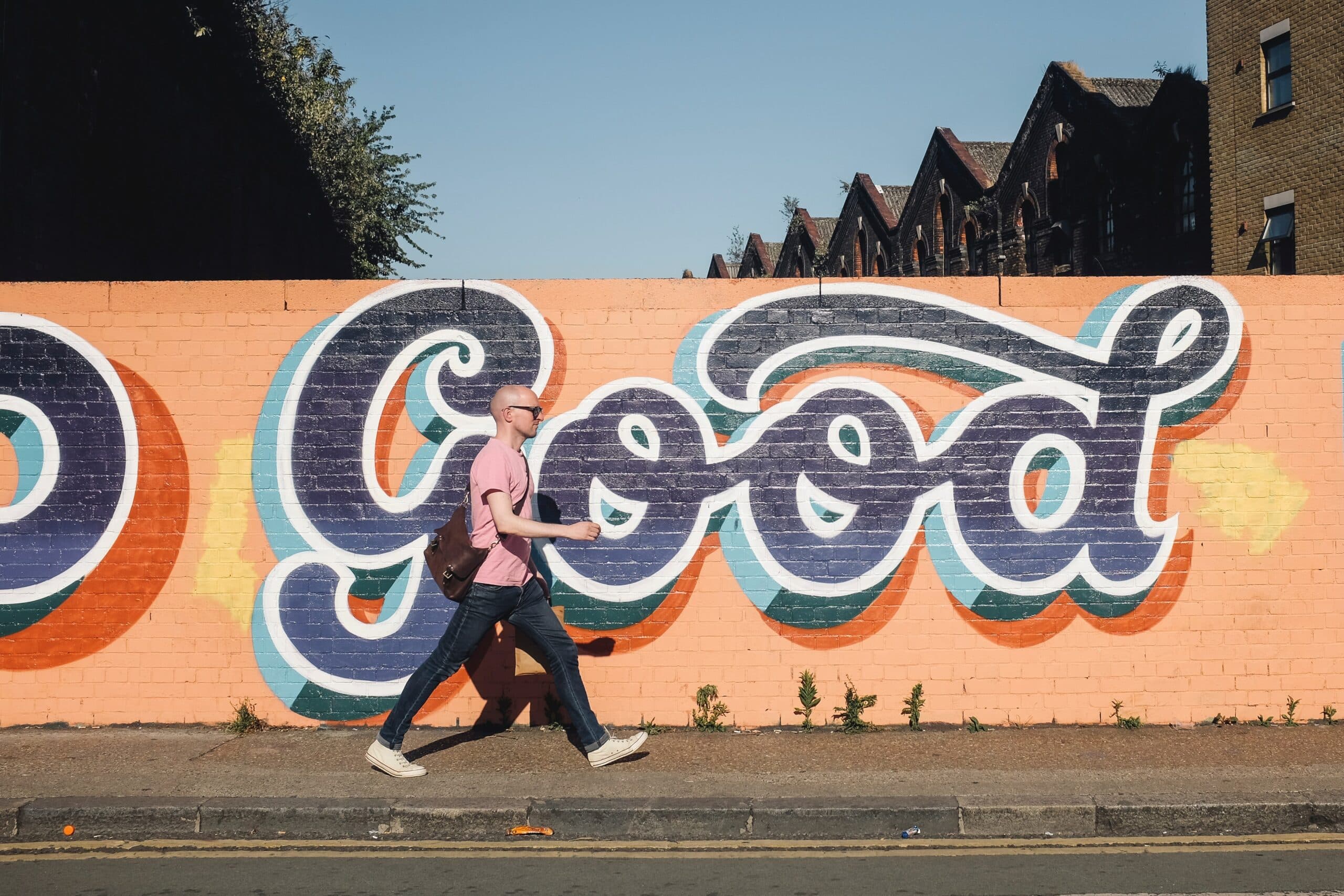 A man wearing a pink shirt and sunglasses walks past a colorful graffiti mural on a wall that says Good. The background includes several buildings with a clear blue sky.