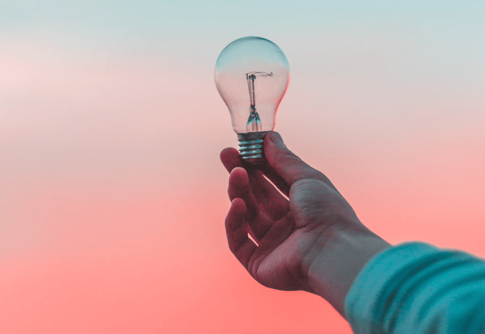 A hand holding a clear light bulb against a blurred background of pink and blue sky, creating a sense of innovation and creativity.