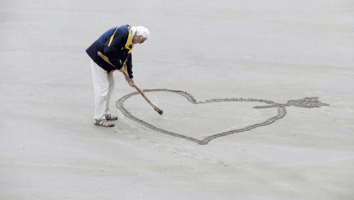 An elderly person in a jacket and light pants draws a large heart with an arrow in the sand using a stick. The beach is mostly empty and overcast, creating a serene atmosphere.