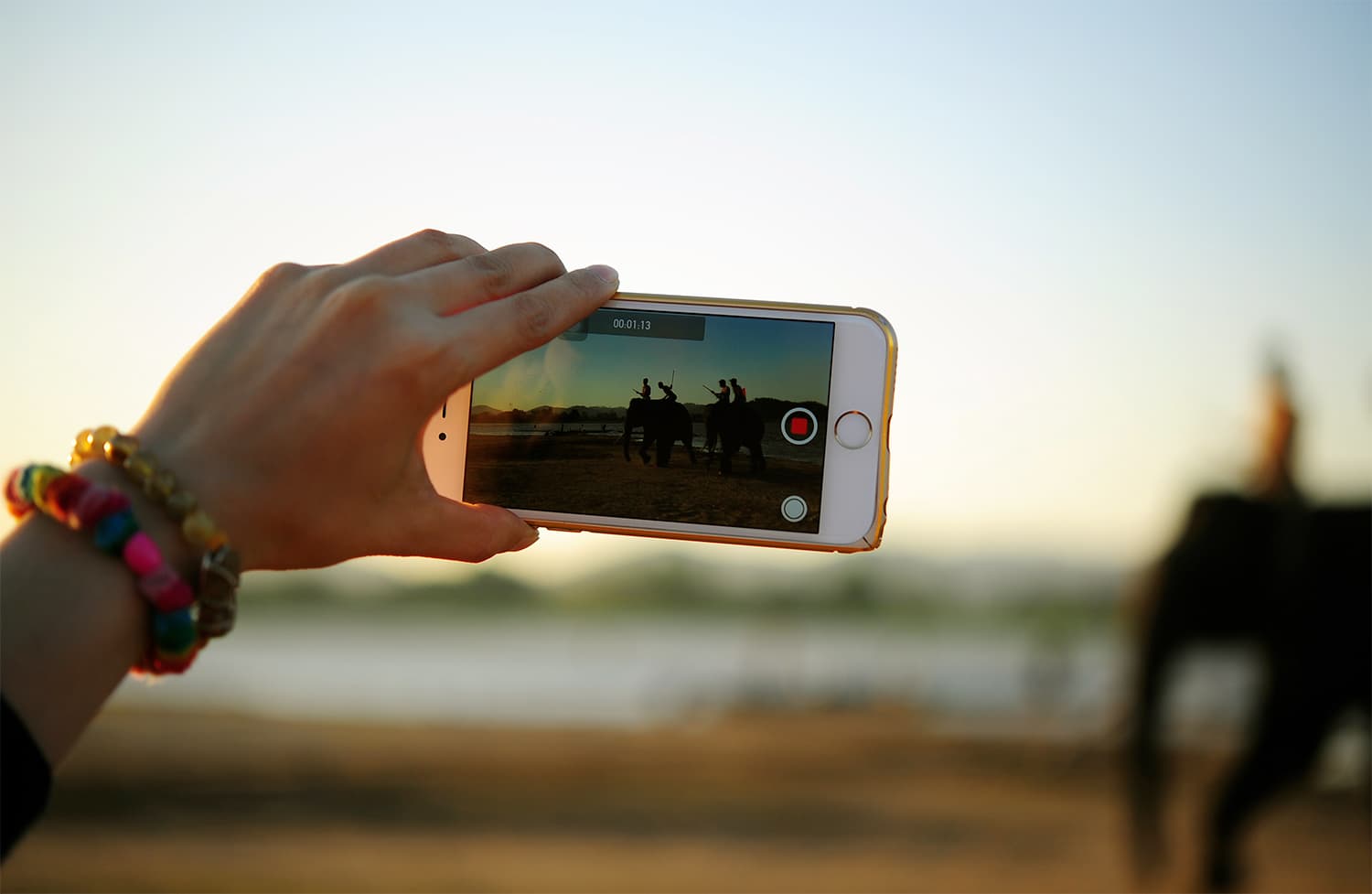 A person holds a smartphone horizontally, capturing a video of a blurred scene with people and a camel against a sunset backdrop. The phone screen shows a recording interface. The person wears colorful bracelets.