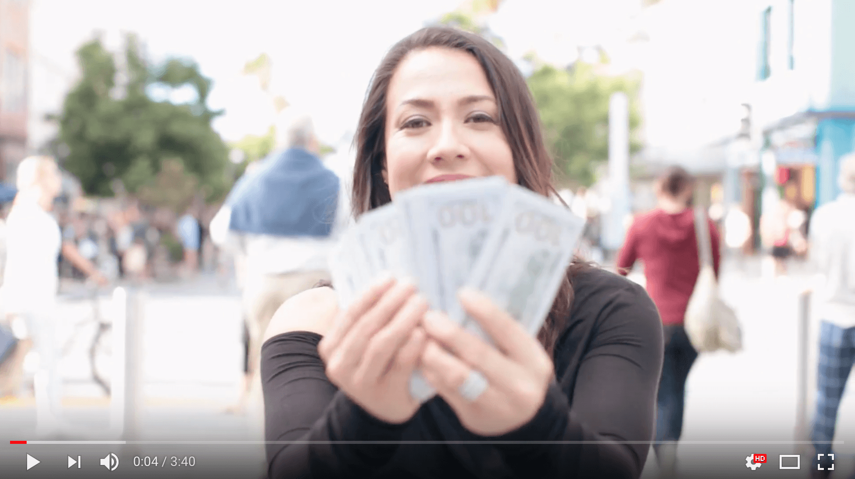 A person holding a fan of 100-dollar bills in both hands, smiling and standing outdoors on a busy street. People and shops are visible in the background, creating a vibrant urban scene.