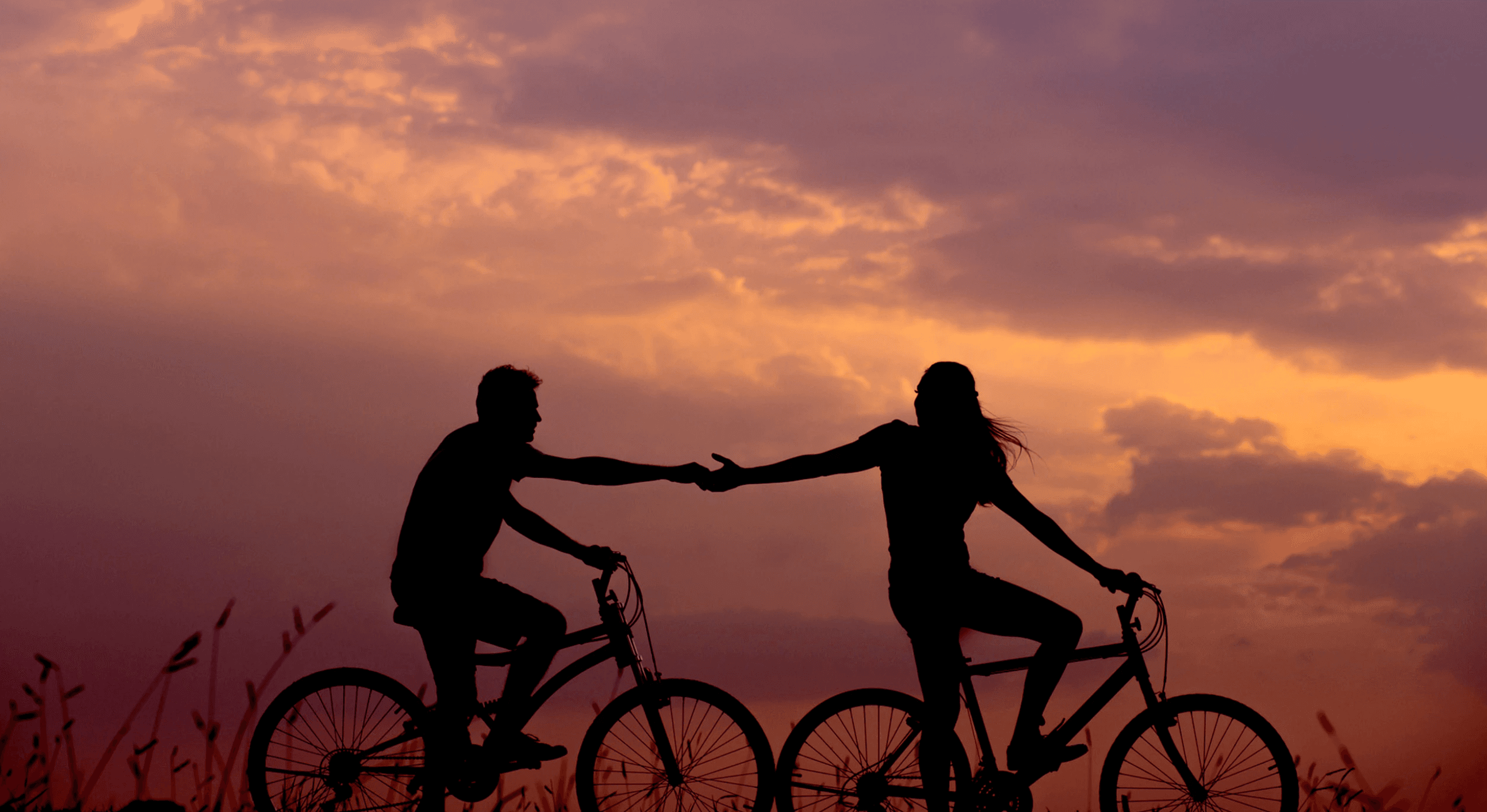 Two people on bicycles silhouetted against a vibrant sunset sky. They are reaching out and holding hands, creating a sense of connection and harmony. The background is filled with clouds tinged with shades of orange and purple.
