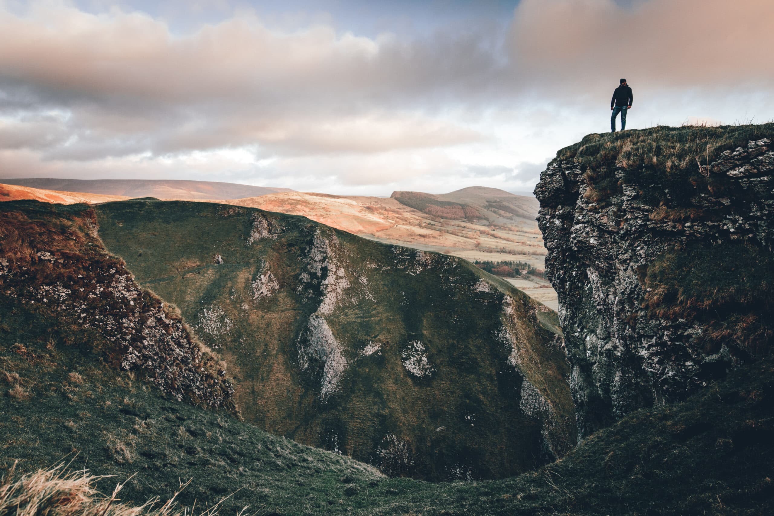 A person stands on the edge of a rugged cliff, overlooking a vast, green valley under a cloudy sky. The landscape features rolling hills and dramatic rock formations, with a warm glow of sunlight illuminating parts of the scene.