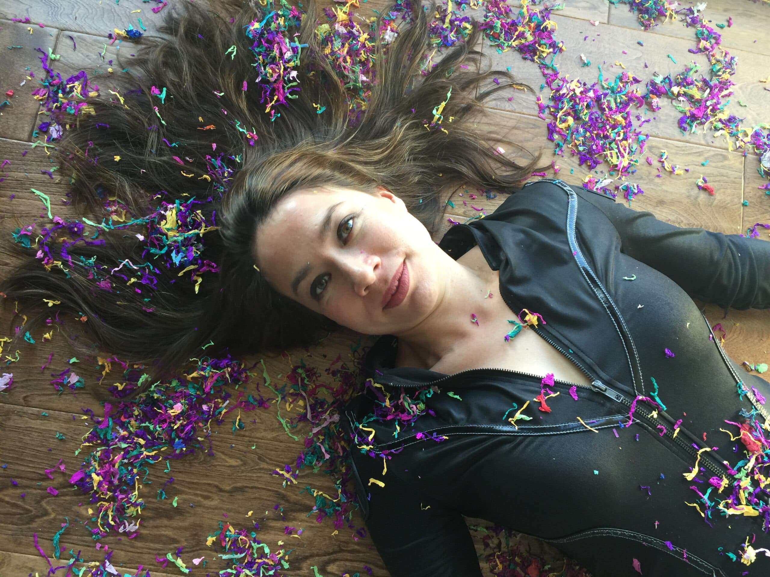 A woman in a black outfit lies on a wooden floor, surrounded by colorful confetti. Her brown hair fans out around her head, and she gazes upwards with a soft smile. The scene conveys a festive, carefree atmosphere.