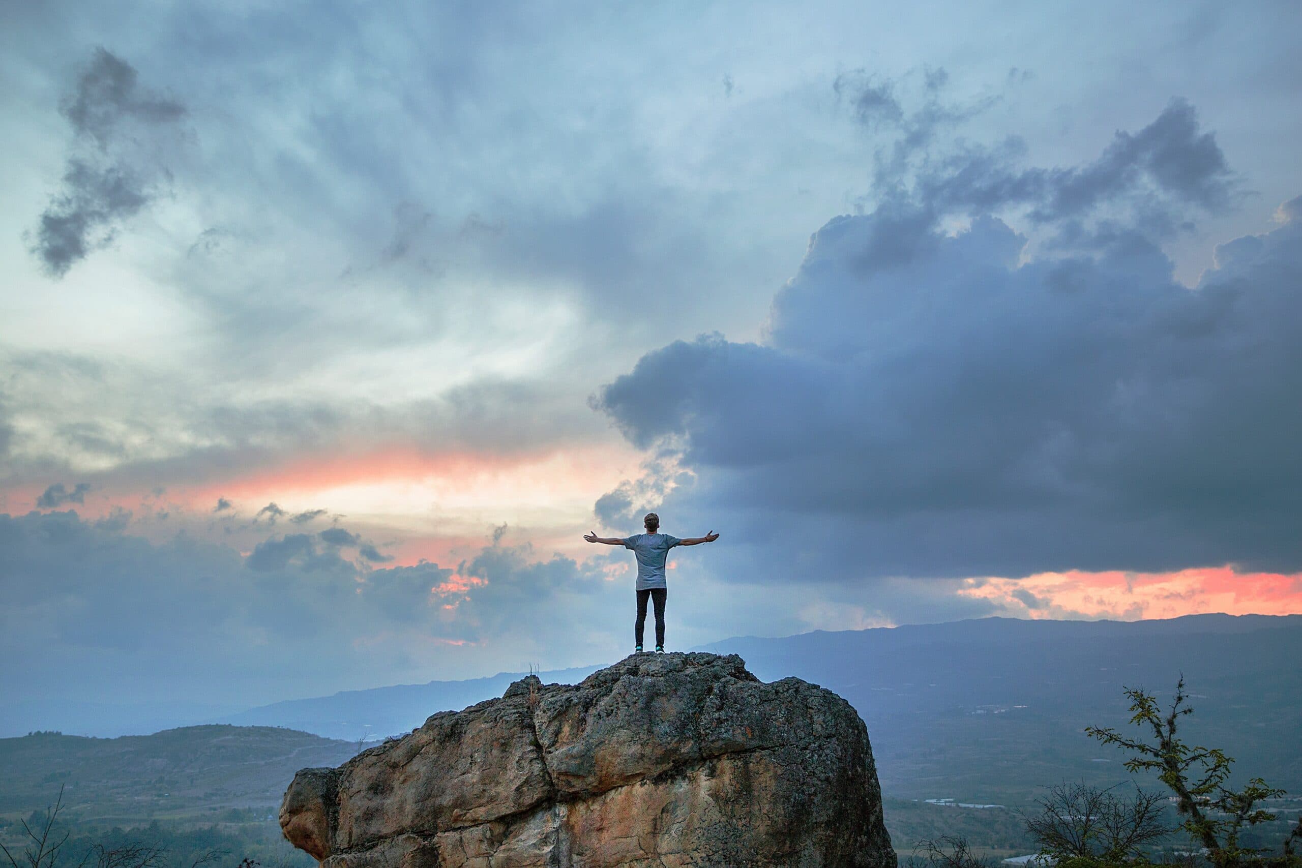 A person stands on a large rock with arms outstretched, silhouetted against a dramatic sunset sky filled with clouds. Rugged terrain stretches into the distance, creating a sense of vastness and freedom.