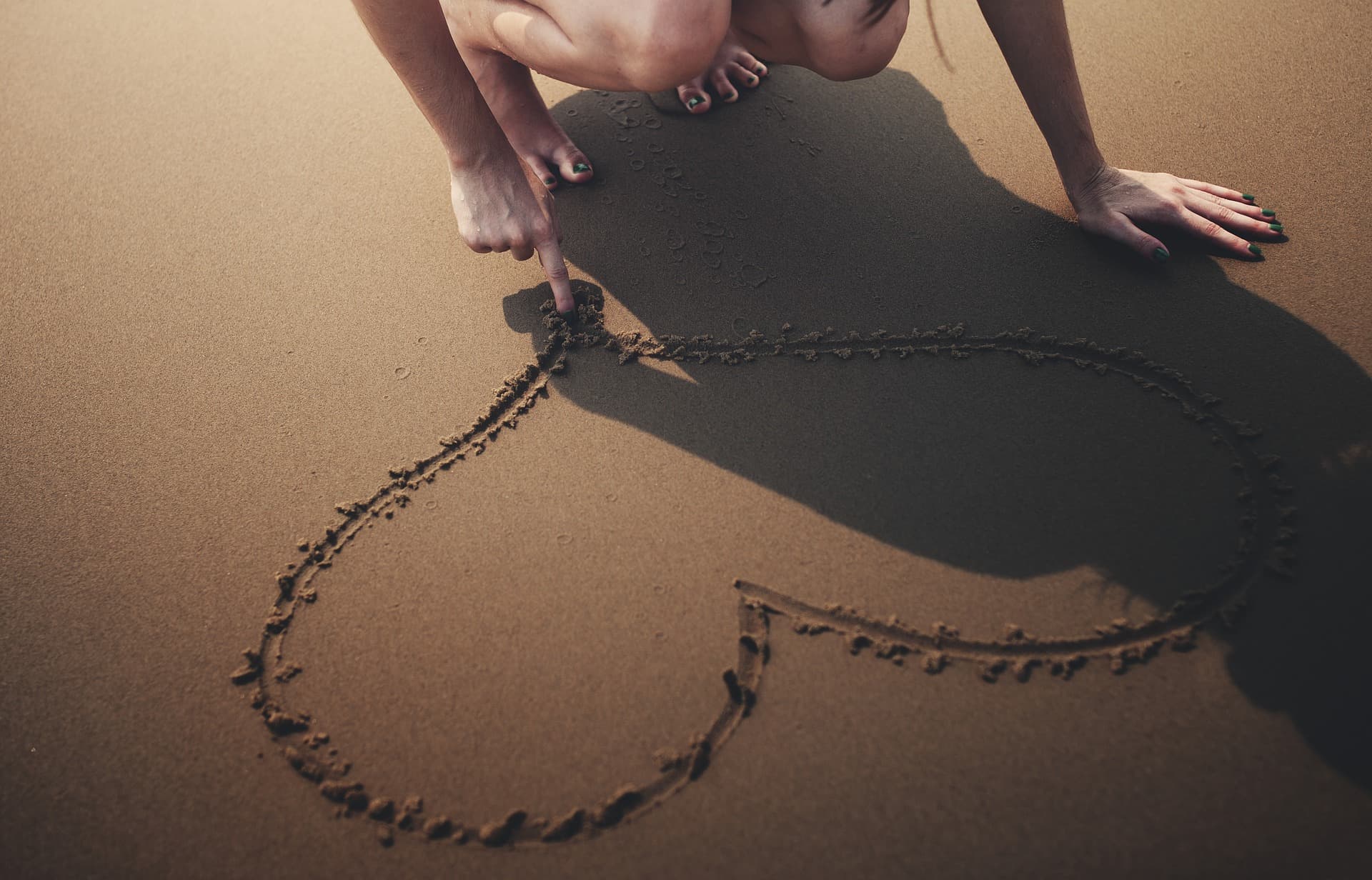 A person drawing a heart shape in the sand with their finger. They are kneeling, and the light shadow is cast beside the heart. The sand has a smooth texture and footprints are barely visible around the edges.
