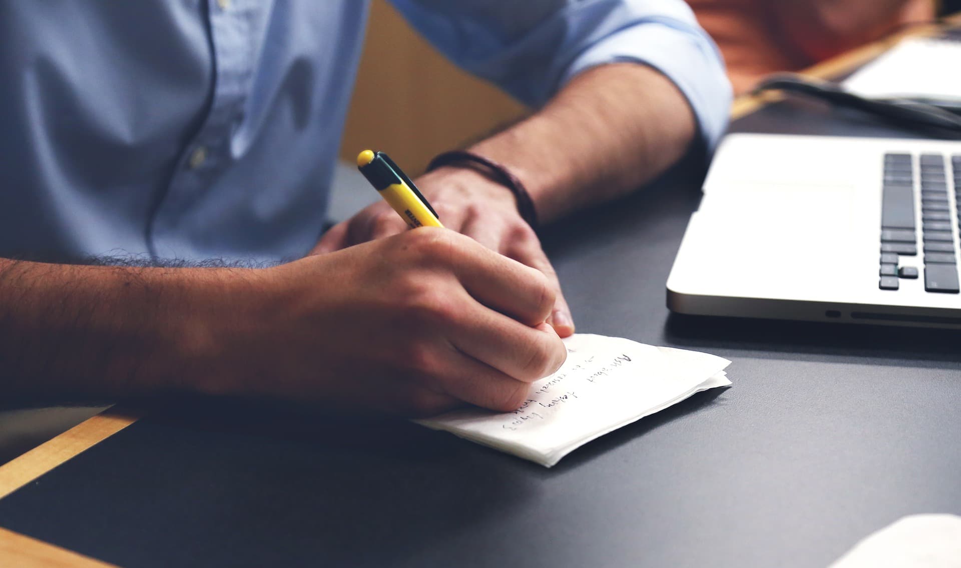 A person writing notes with a pencil on a notepad next to an open laptop on a desk. Theyre wearing a blue shirt, and the focus is on their hand and the notepad.