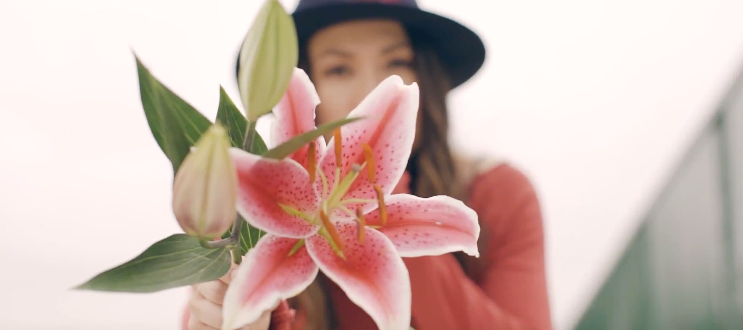 A person in a hat holds a blooming pink lily, partially covering their face. A bud and green leaves accompany the flower, with a blurred background adding depth to the image.