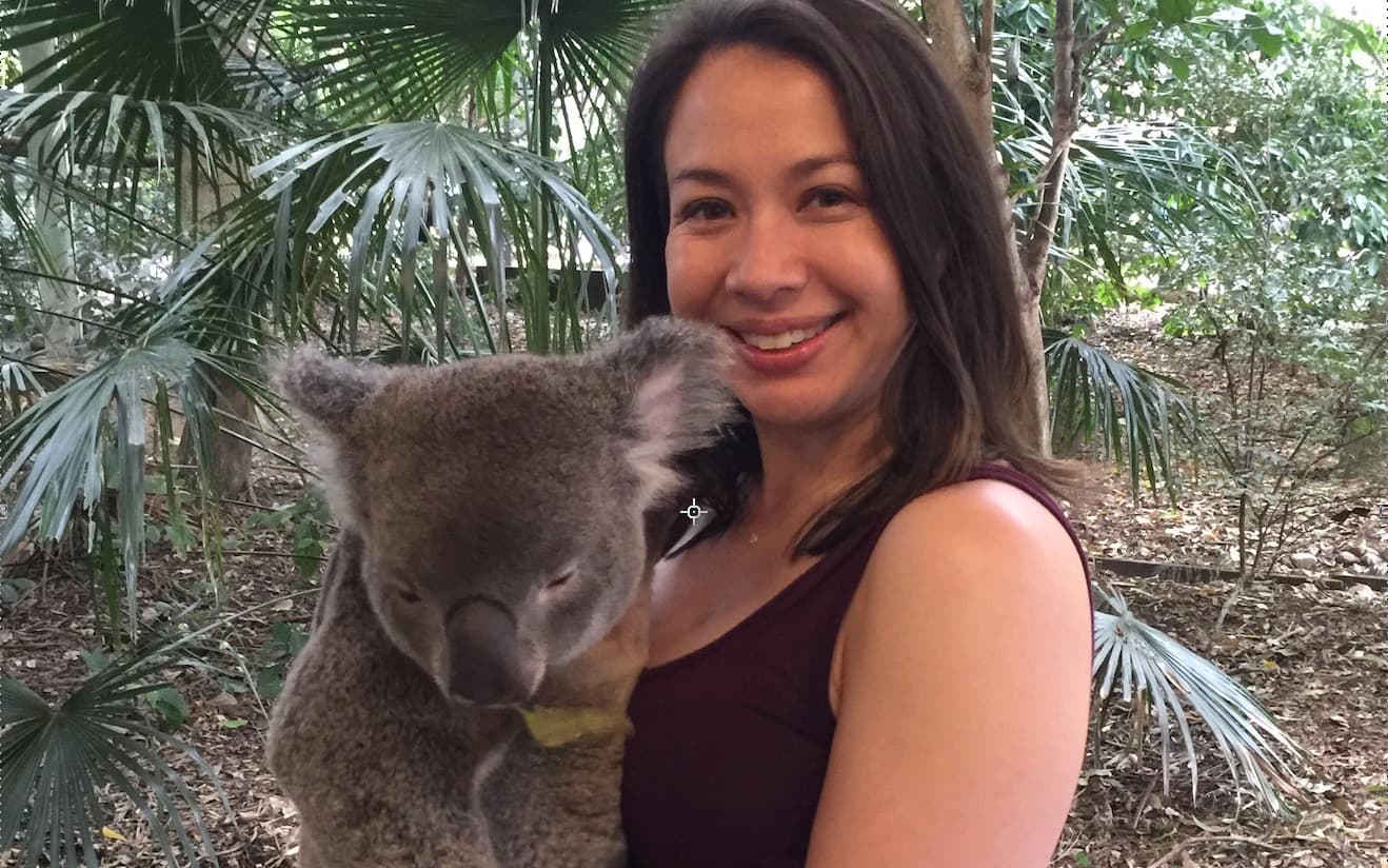 A person smiling while holding a koala in a lush, green outdoor setting with palm leaves and trees in the background.