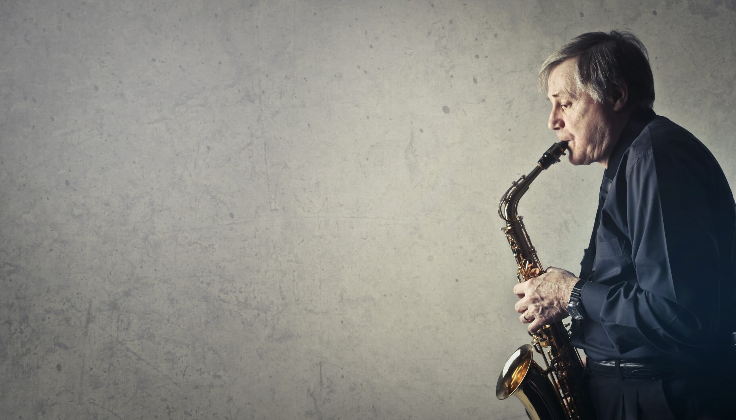 An older man wearing a dark shirt is playing a saxophone. He stands against a textured gray wall, focused on his music. The saxophones golden color contrasts with the neutral background.