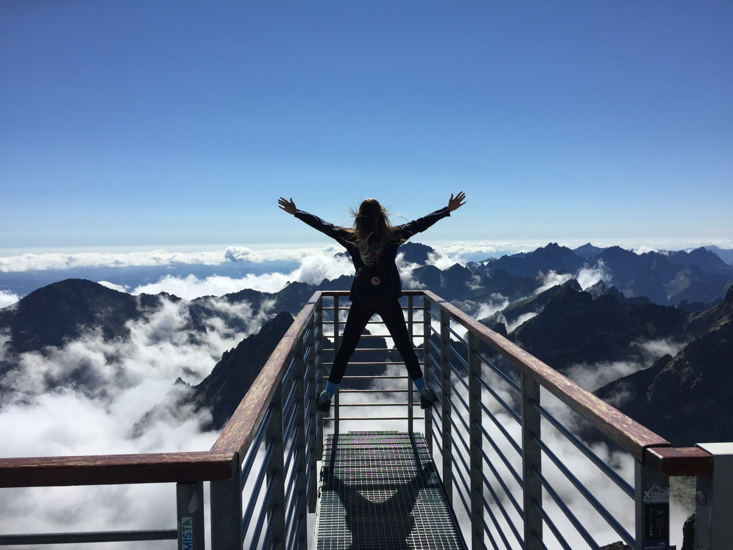 Person with arms outstretched stands on a metal platform overlooking a breathtaking mountain landscape. The sky is clear and the peaks are surrounded by scattered clouds, creating a stunning panoramic view.