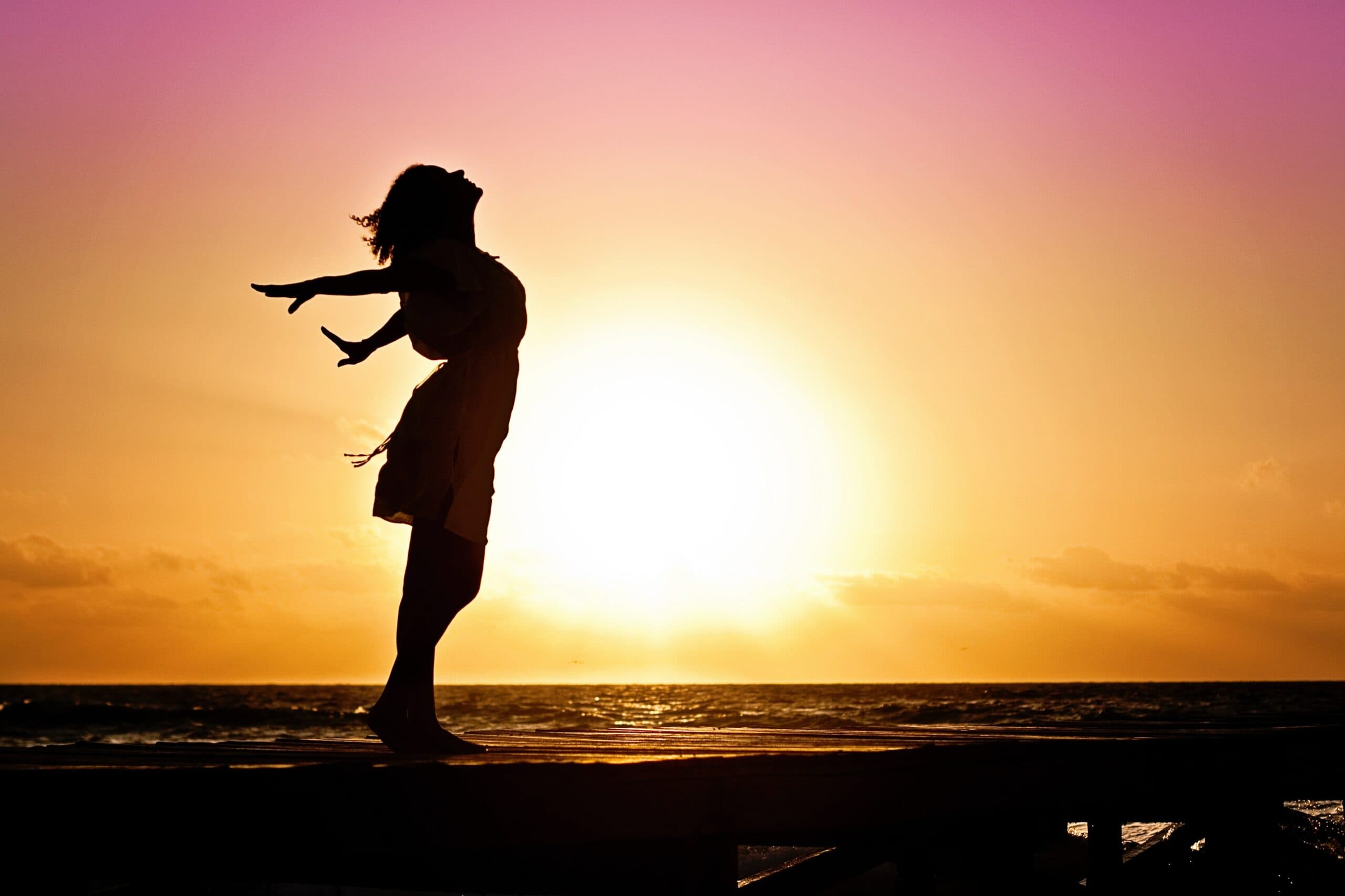 Silhouette of a person standing on a boardwalk, arms outstretched, with a vibrant sunset over the ocean in the background. The sky is a gradient of pink and orange hues.