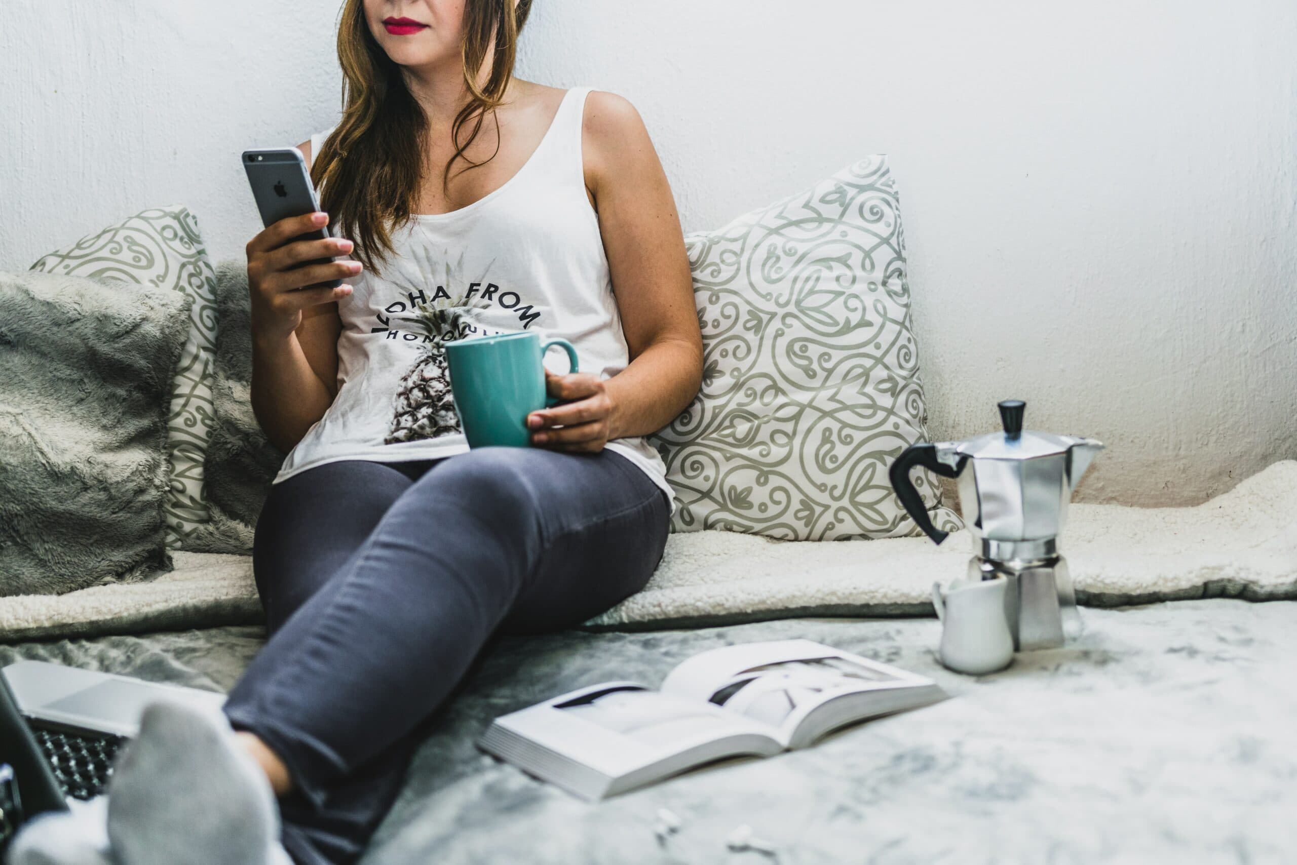 A woman sits comfortably on a sofa, holding a smartphone and a mug. She wears a white tank top and grey pants. An open book and a coffee maker are on the sofa beside her. The setting is casual and relaxed.