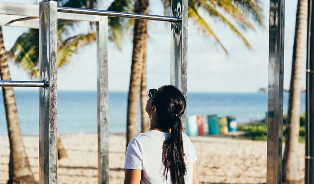A woman with long dark hair stands by metal exercise bars on a sandy beach, with palm trees and the ocean in the background. She is wearing a white T-shirt and sunglasses, gazing at the equipment.
