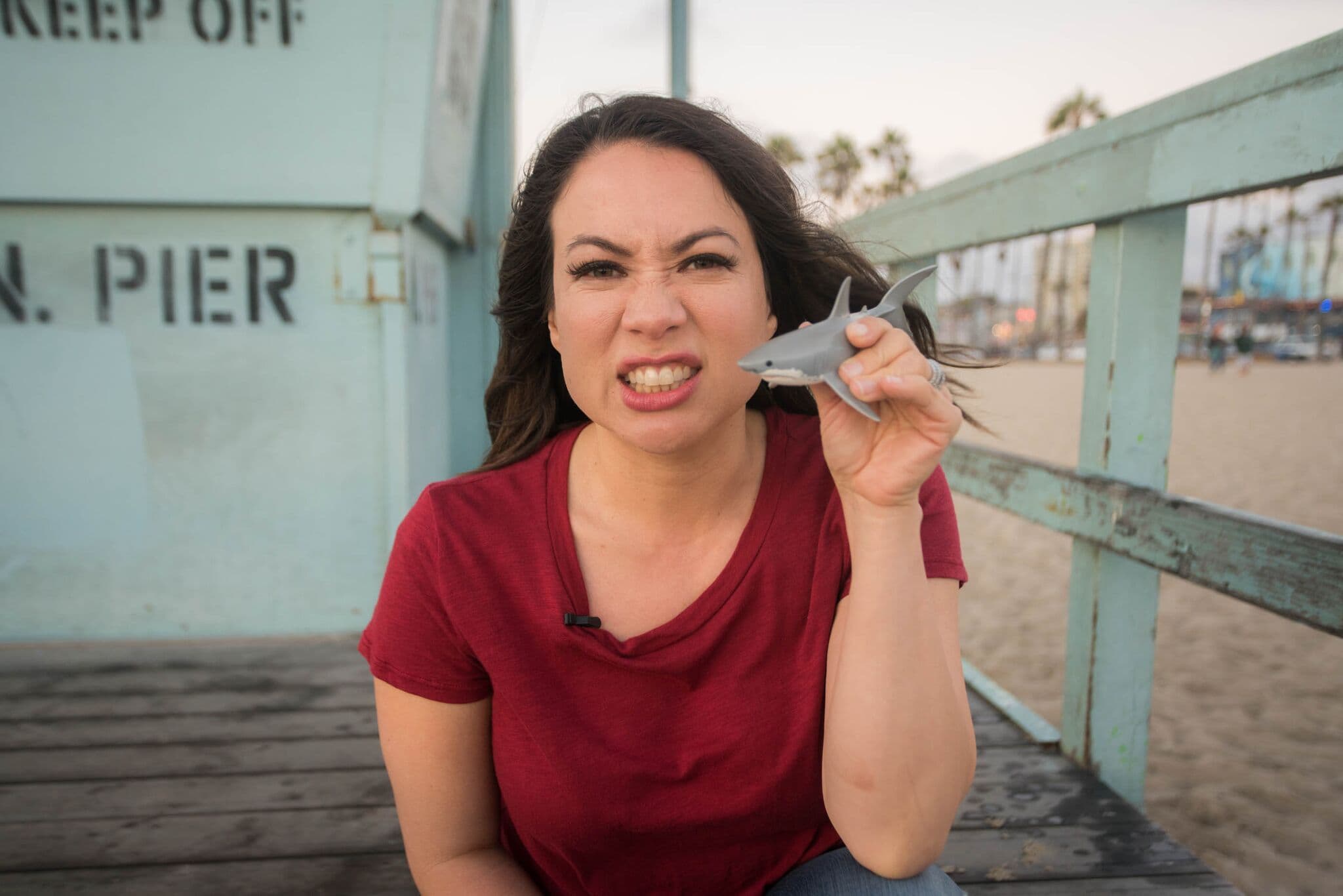 A woman playfully holding a toy shark in her teeth, sitting on a wooden boardwalk at the beach. She wears a red shirt, and the background shows lifeguard signage and palm trees.
