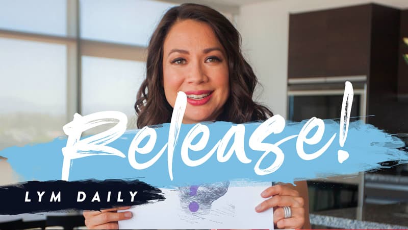 A woman with long dark hair smiles and holds a card with a purple question mark. The word Release! is displayed prominently in white, overlaid on a blue brushstroke. The text LYM Daily appears in a dark band at the bottom left.