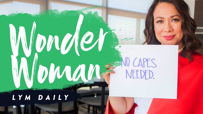 A woman wearing a red outfit holds a sign that reads, No capes needed. The left side features large text saying Wonder Woman with LYM Daily below it. The background shows a bright room with large windows.