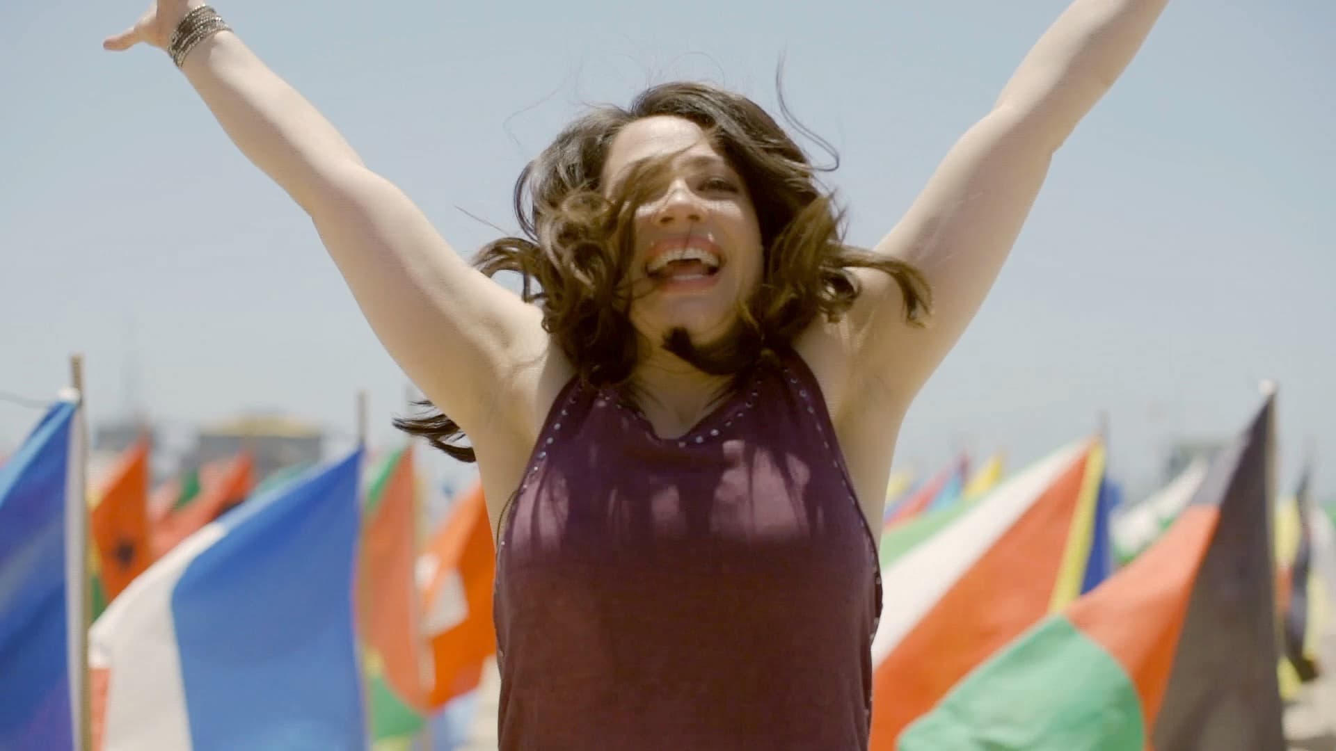 A person with brown hair and a purple sleeveless top joyfully raises their arms amid a colorful display of international flags, set against a clear blue sky.