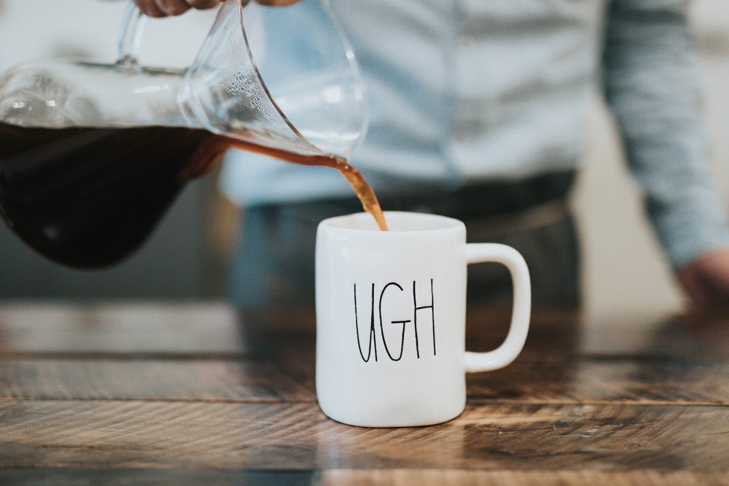 A person pours coffee from a glass carafe into a white mug labeled UGH on a wooden table. The background is blurred, focusing on the coffee being poured.