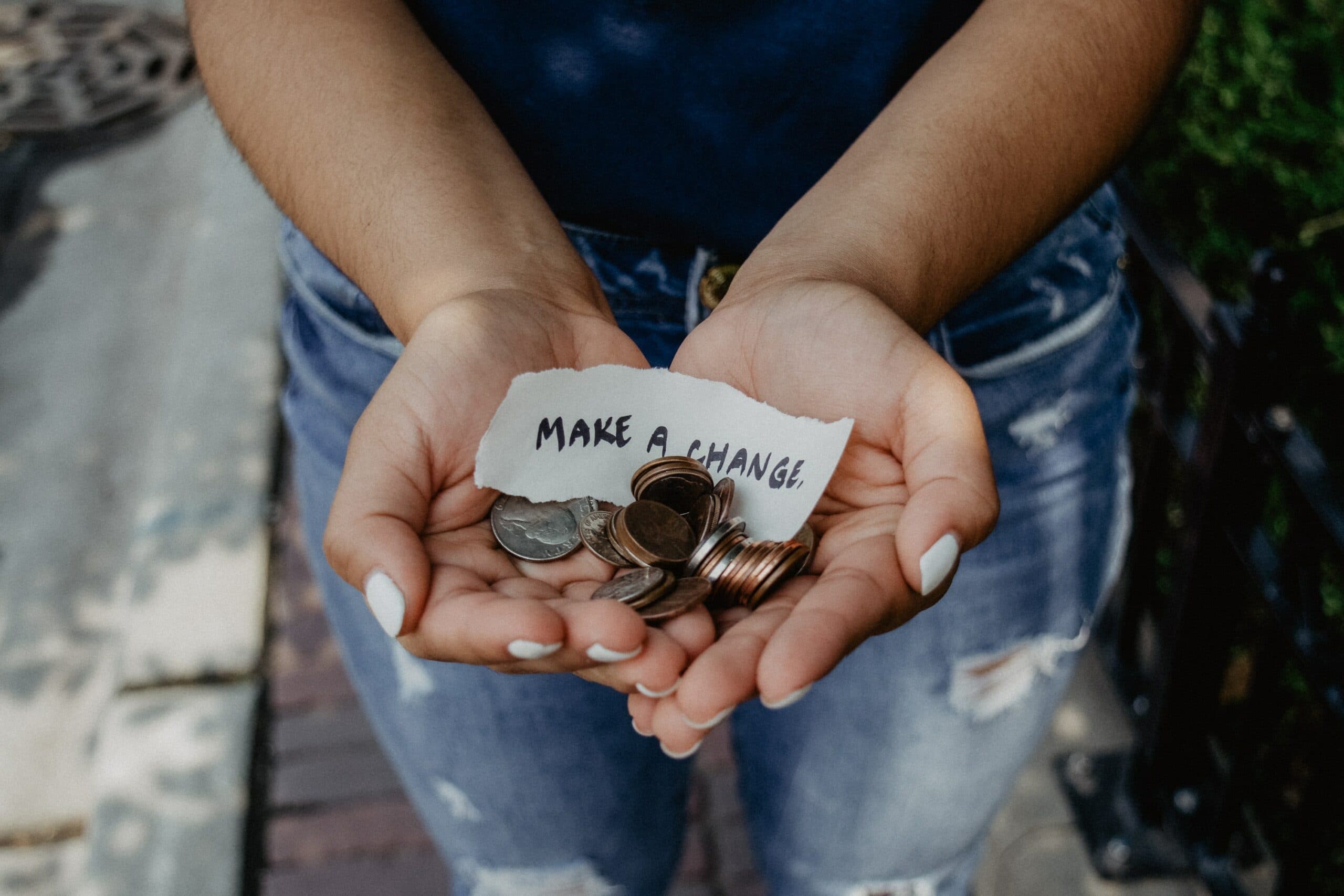 A person with light-painted nails holds a handful of coins and a small paper note that reads MAKE A CHANGE. The background is slightly blurred, featuring a brick path and greenery.