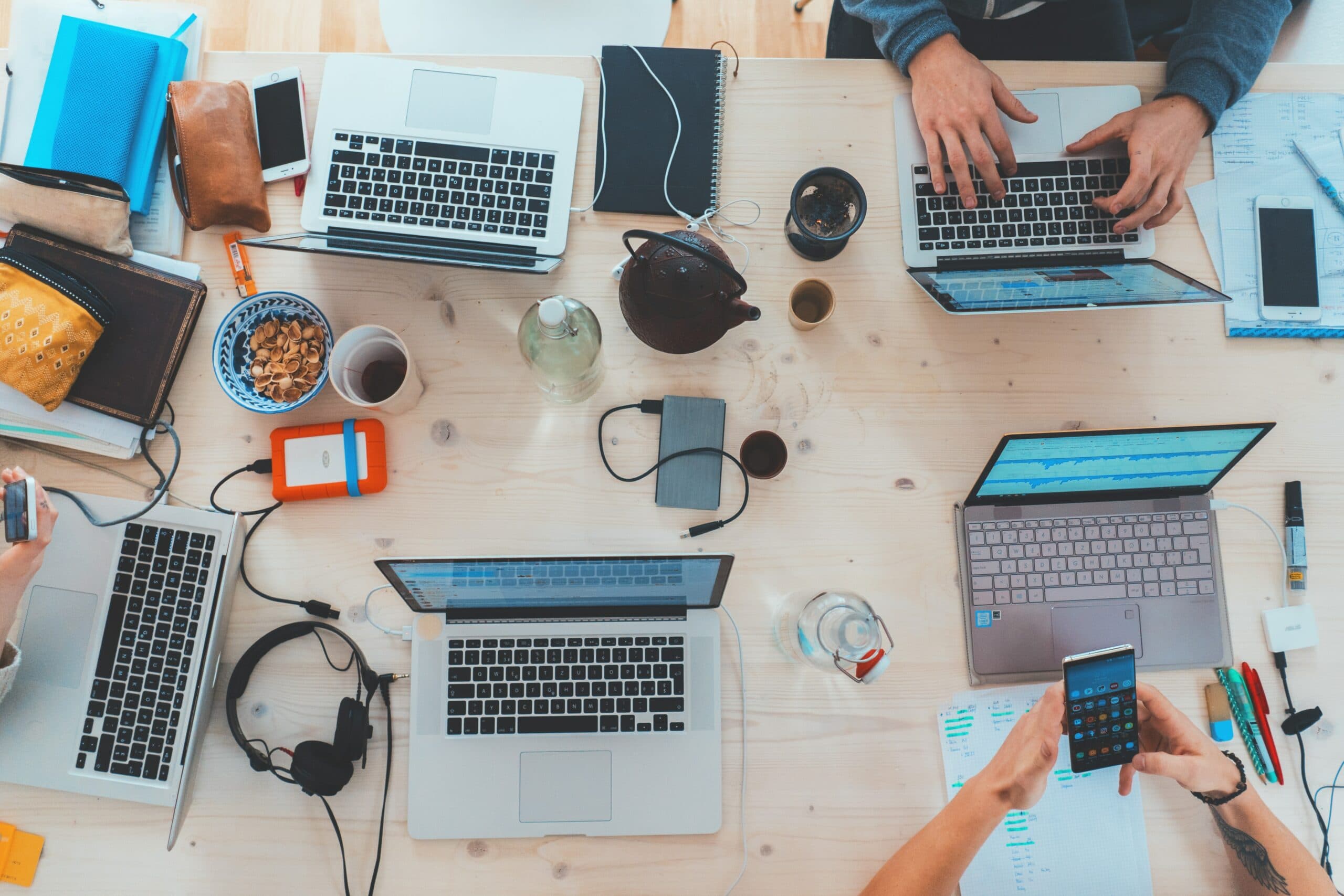 People are working at a desk with several laptops, phones, notebooks, and a tablet. Snacks, headphones, a teapot, and a camera are also on the table, creating a busy, collaborative work environment.