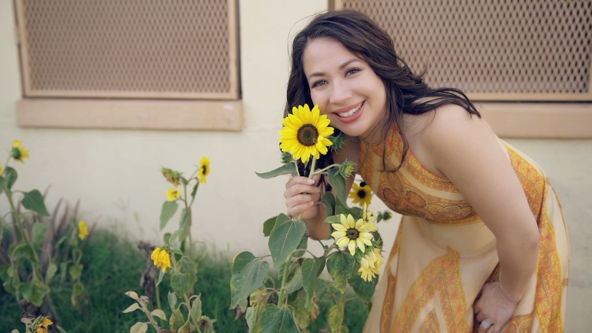 A woman in a yellow dress leans toward blooming sunflowers, smiling at the camera. She is outdoors in front of a building with lattice-patterned windows. The sunflowers add a touch of vibrant color to the scene.