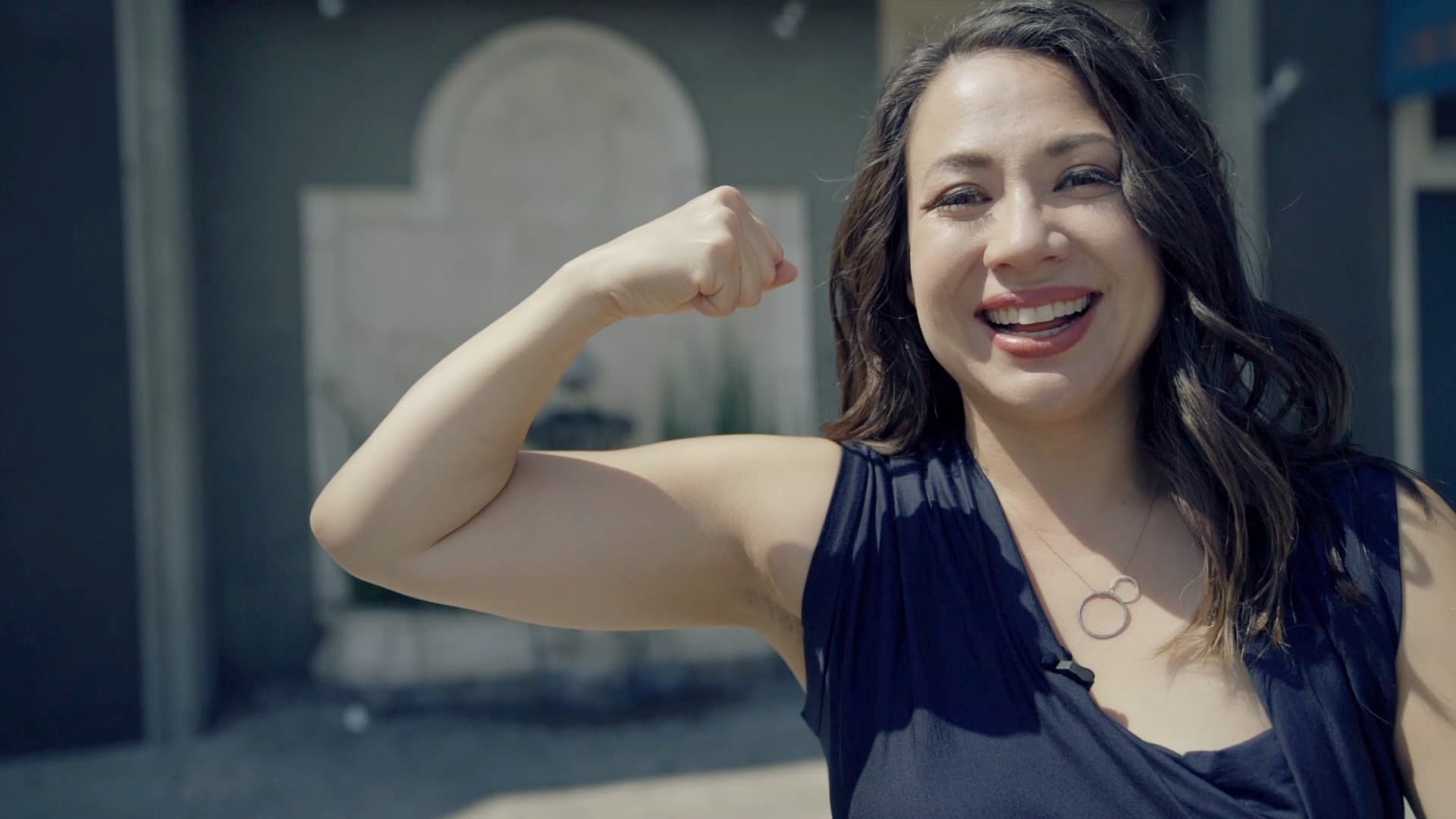 A woman smiling and flexing her arm in a confident pose outside, with a blurred building in the background. Shes wearing a navy top and a circular pendant necklace, standing under a clear sky.