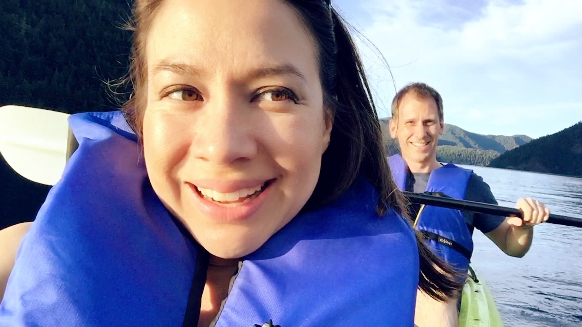 A woman and a man, both wearing blue life jackets, are kayaking on a lake. The woman is smiling at the camera, while the man, slightly behind her, is also smiling. The background features calm water and green hills under a blue sky.