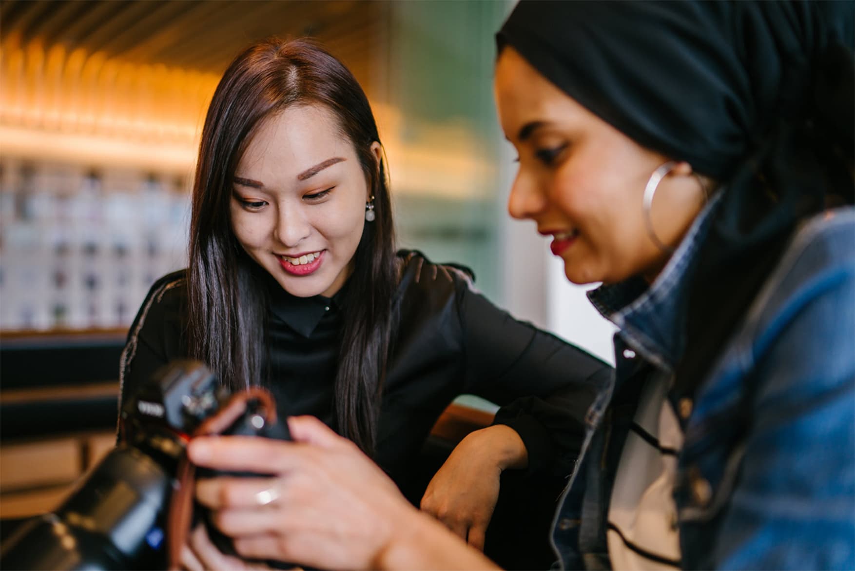 Two women are sitting closely and smiling while looking at images on a camera. One woman is wearing a black shirt and the other a denim jacket and headscarf. They appear to be in a warmly lit indoor space.