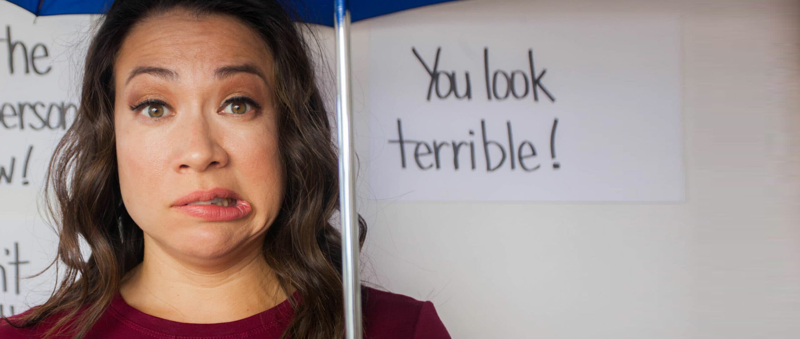 A woman under an umbrella makes a grimacing face. Behind her, a partially visible sign reads You look terrible! in bold letters.