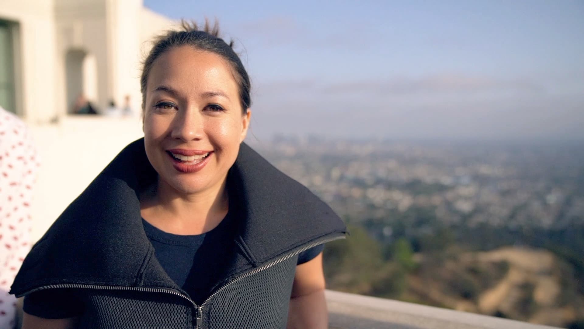 A woman stands smiling with a view of a cityscape in the background. She is wearing a black top with a large collar and has her hair pulled back. The scene appears to be taken on a clear, sunny day.