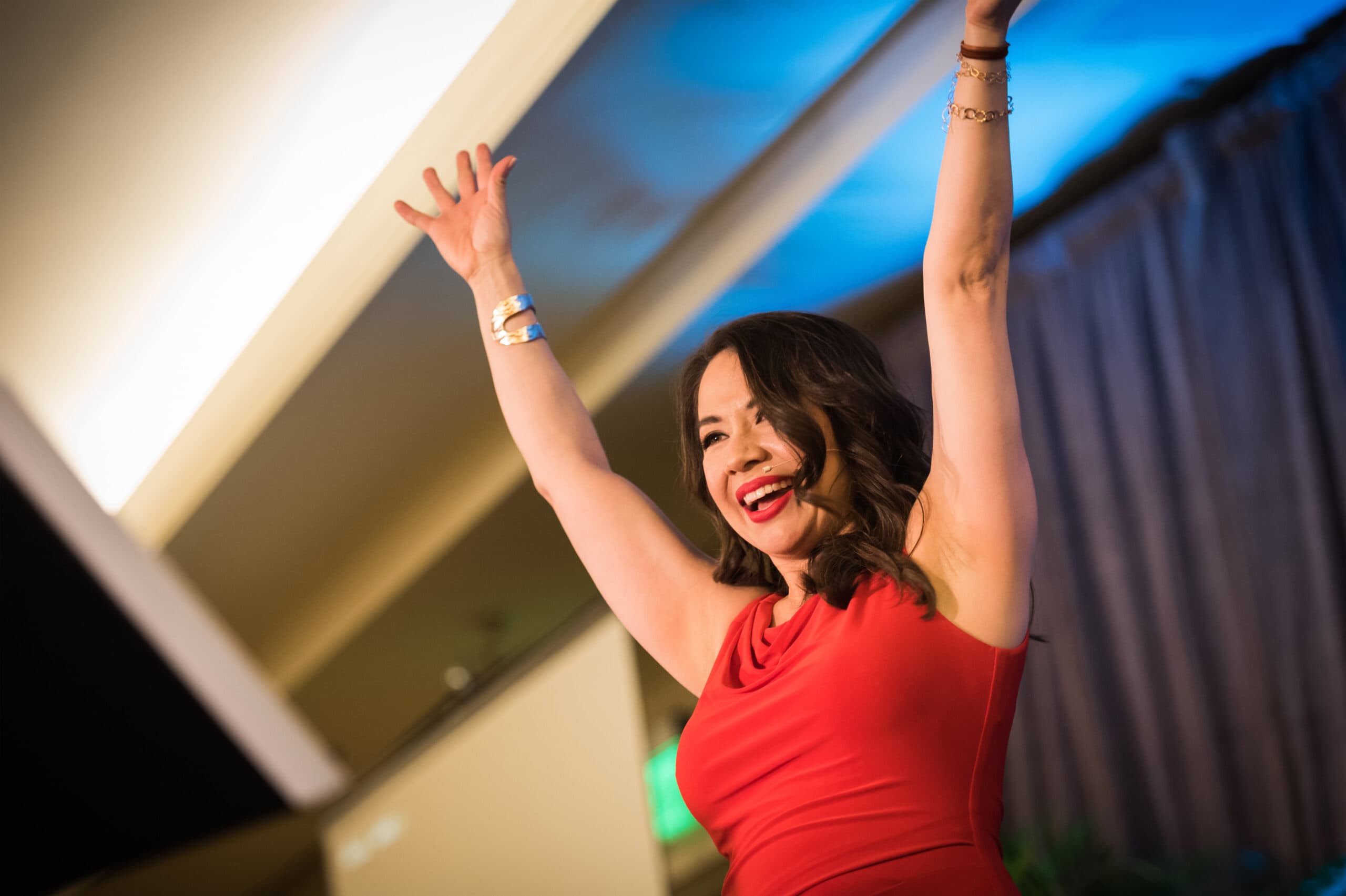 A woman in a red dress is smiling and raising her arms enthusiastically. She stands in a room with a curtain and soft lighting, exuding a sense of excitement and joy.