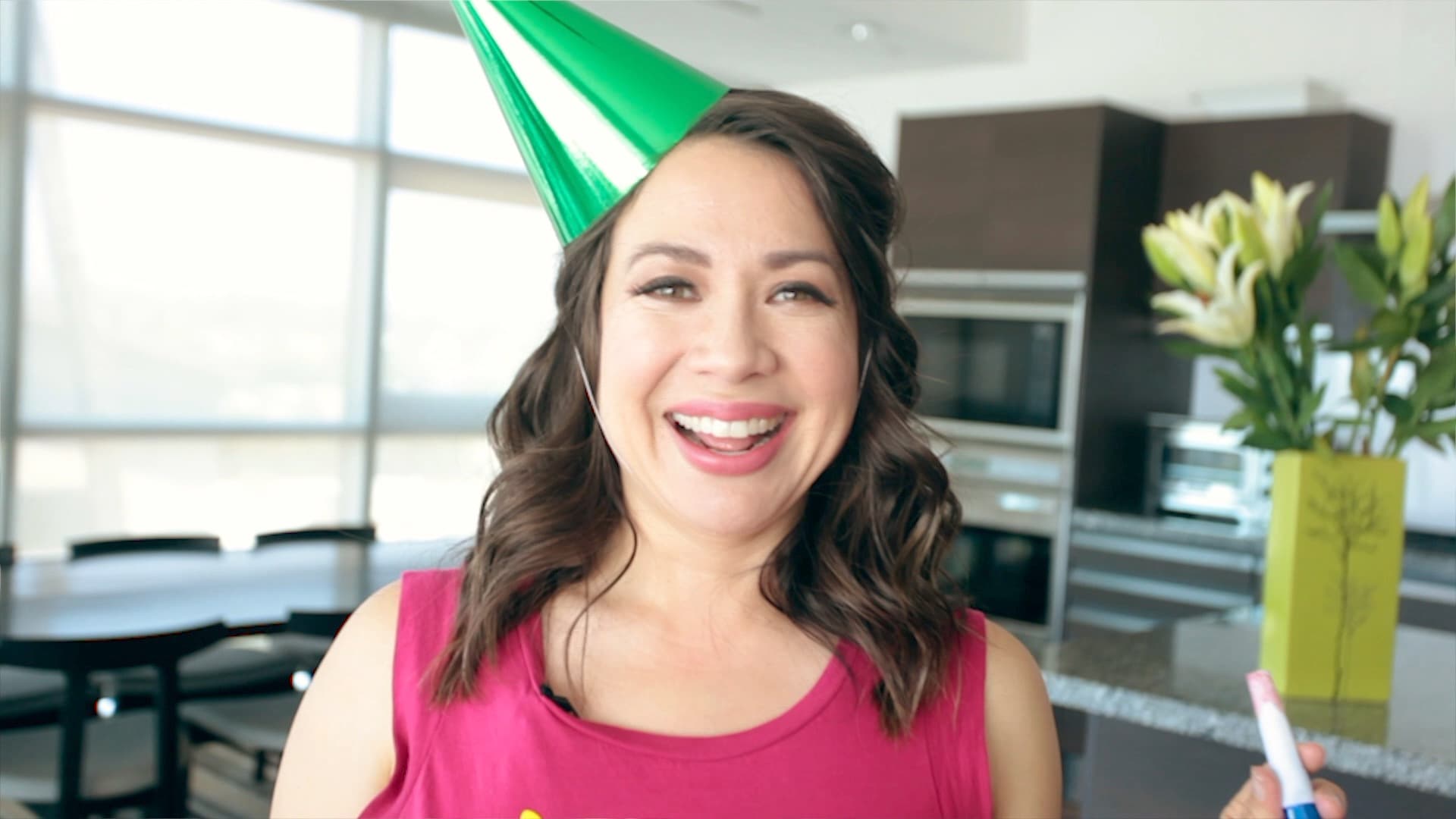 A woman in a pink shirt smiles while wearing a shiny green party hat. She stands in a modern kitchen with large windows and holds a colorful noise maker. In the background, there are white lilies in a bright yellow vase.