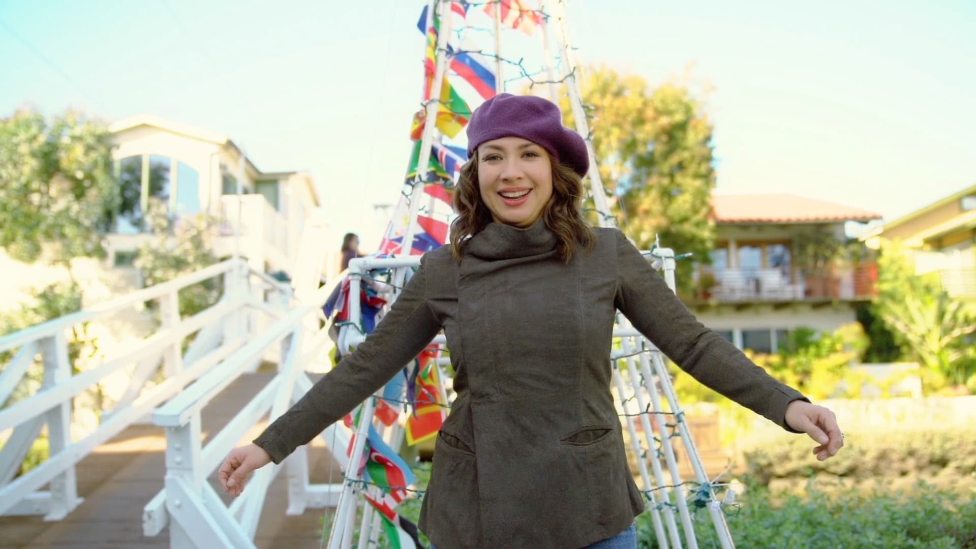Woman in a purple beret and green jacket smiles with arms outstretched in front of a bridge adorned with colorful flags. Bright houses and greenery are visible in the background under a clear blue sky.