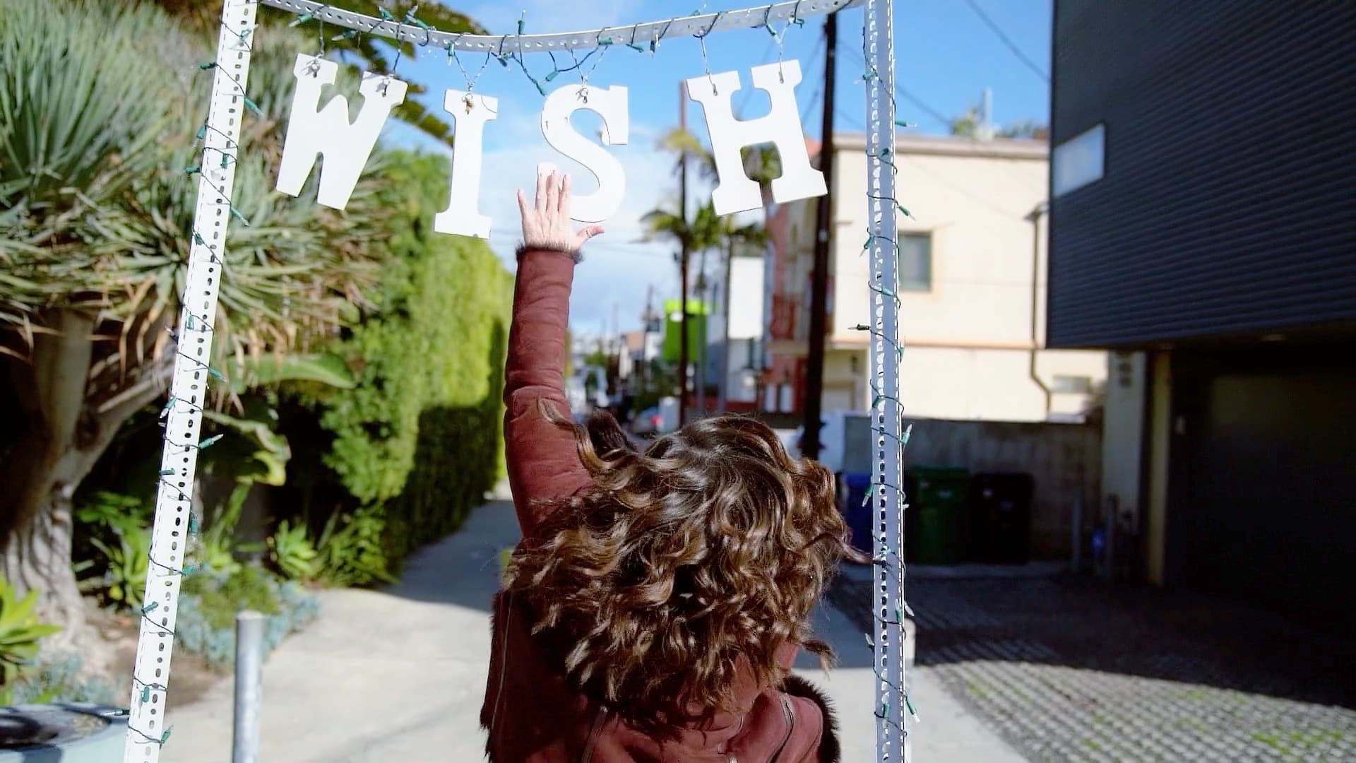 A person with curly hair and a brown coat reaches up to touch a sign that reads WISH hanging on a metal frame. The scene is set outdoors in a narrow, sunny alley lined with greenery and buildings.