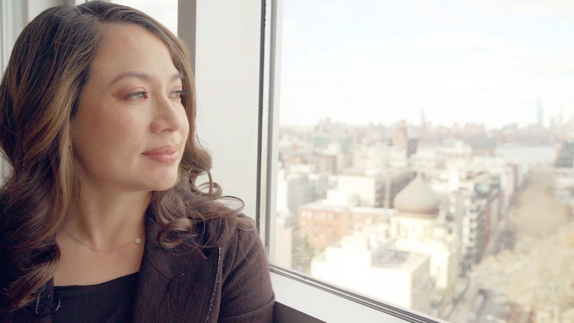 A woman with long, wavy hair sits by a window, gazing outside with a thoughtful expression. The view includes a cityscape with buildings under a cloudy sky. She wears a dark top and a subtle necklace.