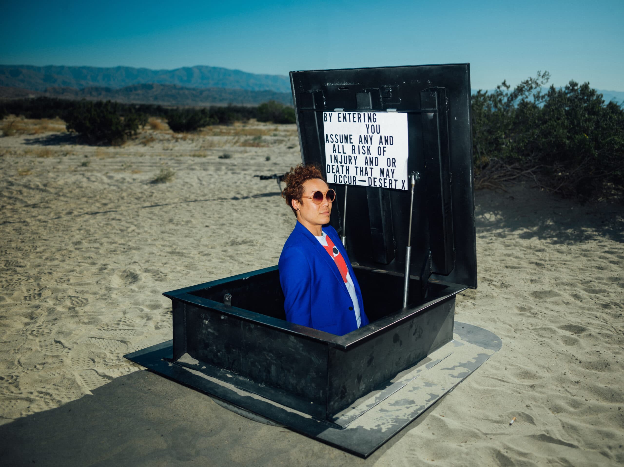 A person wearing a blue jacket and sunglasses stands inside an open hatch in a desert setting. A sign on the hatch reads, BY ENTERING YOU ASSUME ANY AND ALL RISK OF INJURY AND OR DEATH THAT MAY OCCUR - DESERT. Shrubs and mountains are visible in the background.