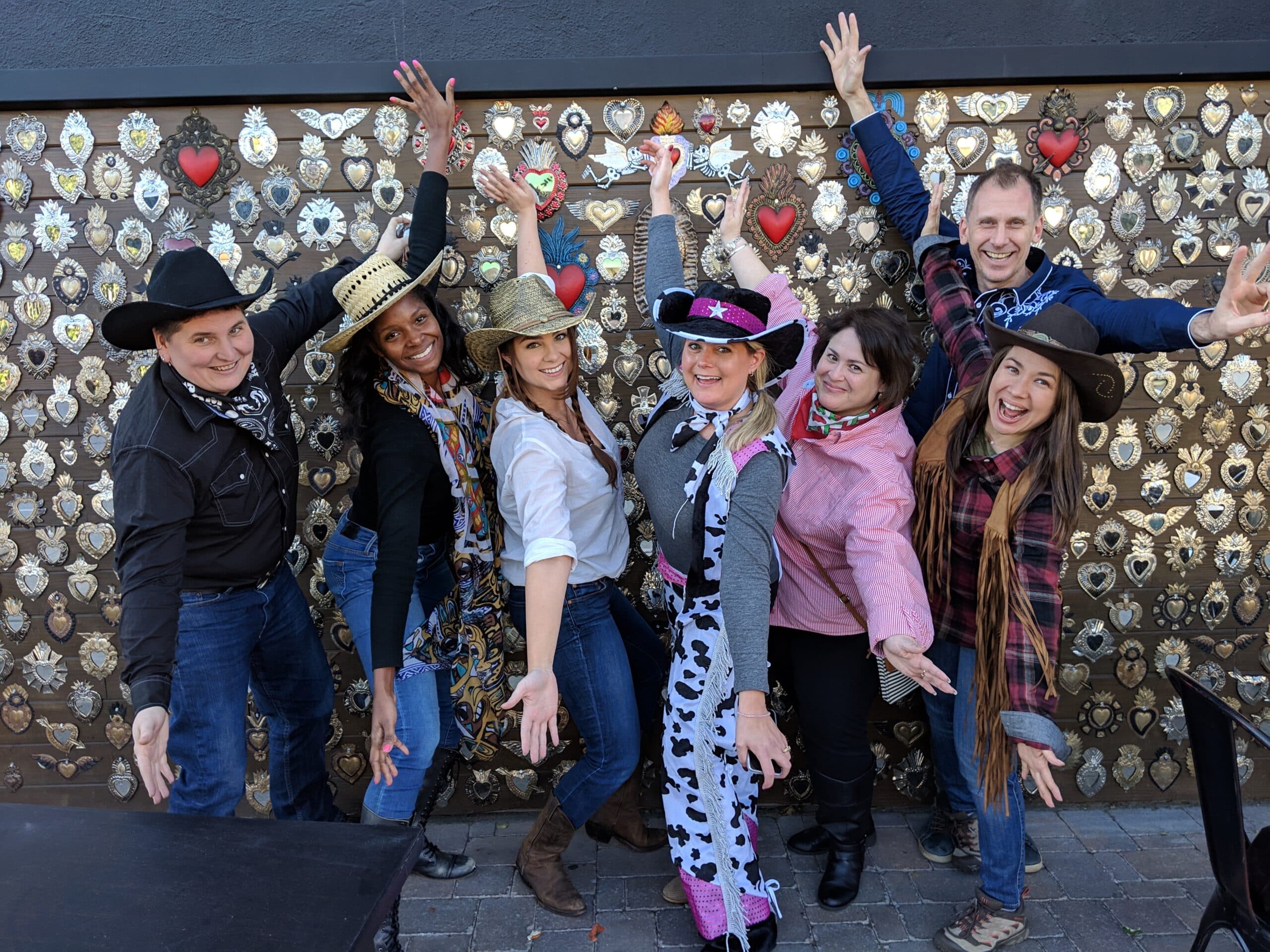 A group of seven people pose playfully in cowboy hats and colorful outfits against a wall decorated with badges and hearts. They are smiling and gesturing with their arms outstretched, capturing a fun moment.