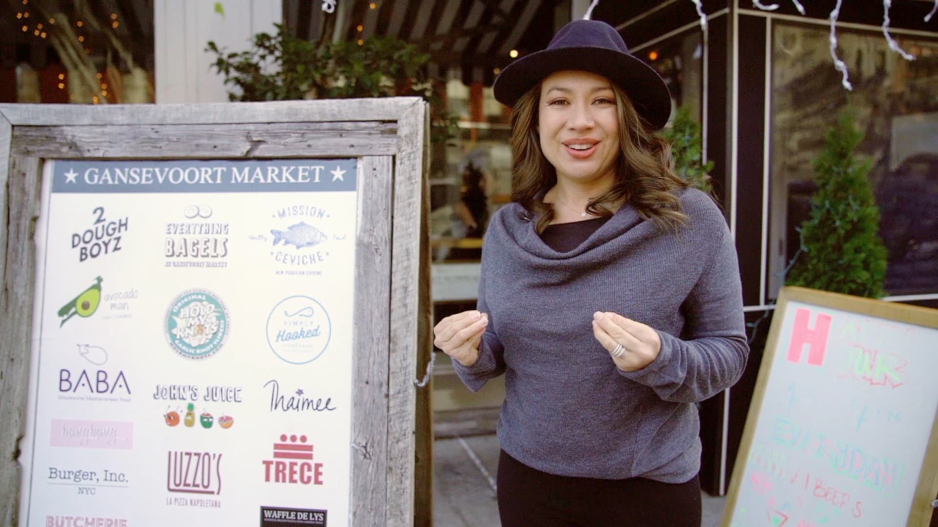 A person wearing a dark hat and gray sweater stands beside a wooden sign at Gansevoort Market. The sign displays various vendor logos, including Dough Boyz, Everything Bagels, and Trece. Festive lights and a chalkboard sign are in the background.
