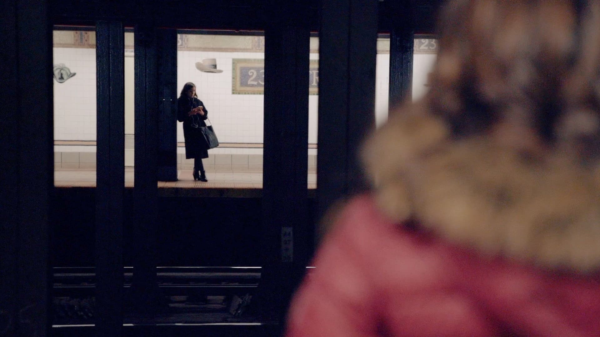A subway platform scene shows a woman in a black coat waiting across the tracks, while another person stands in the foreground wearing a red jacket with a fur-lined hood. Subway signs and tiled walls are visible in the background.