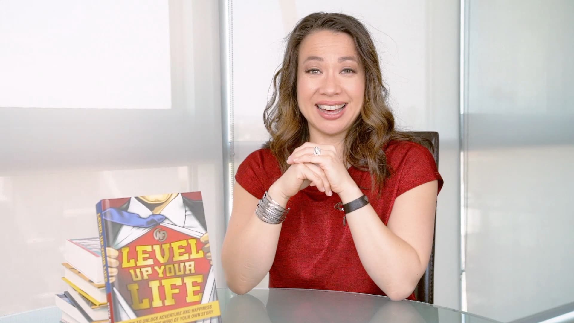 A woman with long brown hair, wearing a red shirt, sits at a glass table. She is smiling with hands clasped. A book titled Level Up Your Life with a superhero cover design is displayed on the table.