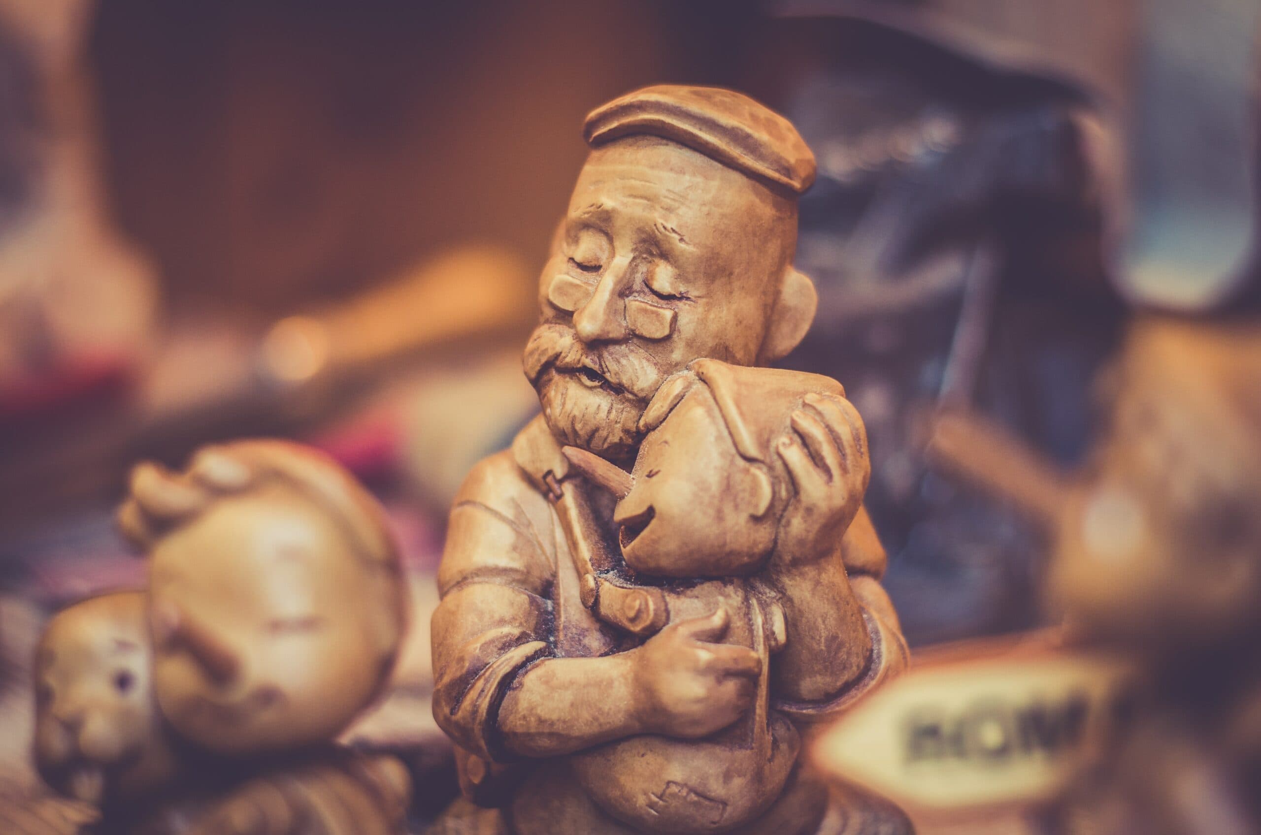 A carved wooden sculpture depicting a smiling elderly man with a cap, gently holding a small child close. Both figures have their eyes closed, conveying a sense of warmth and affection.其他 objects are blurred in the background.