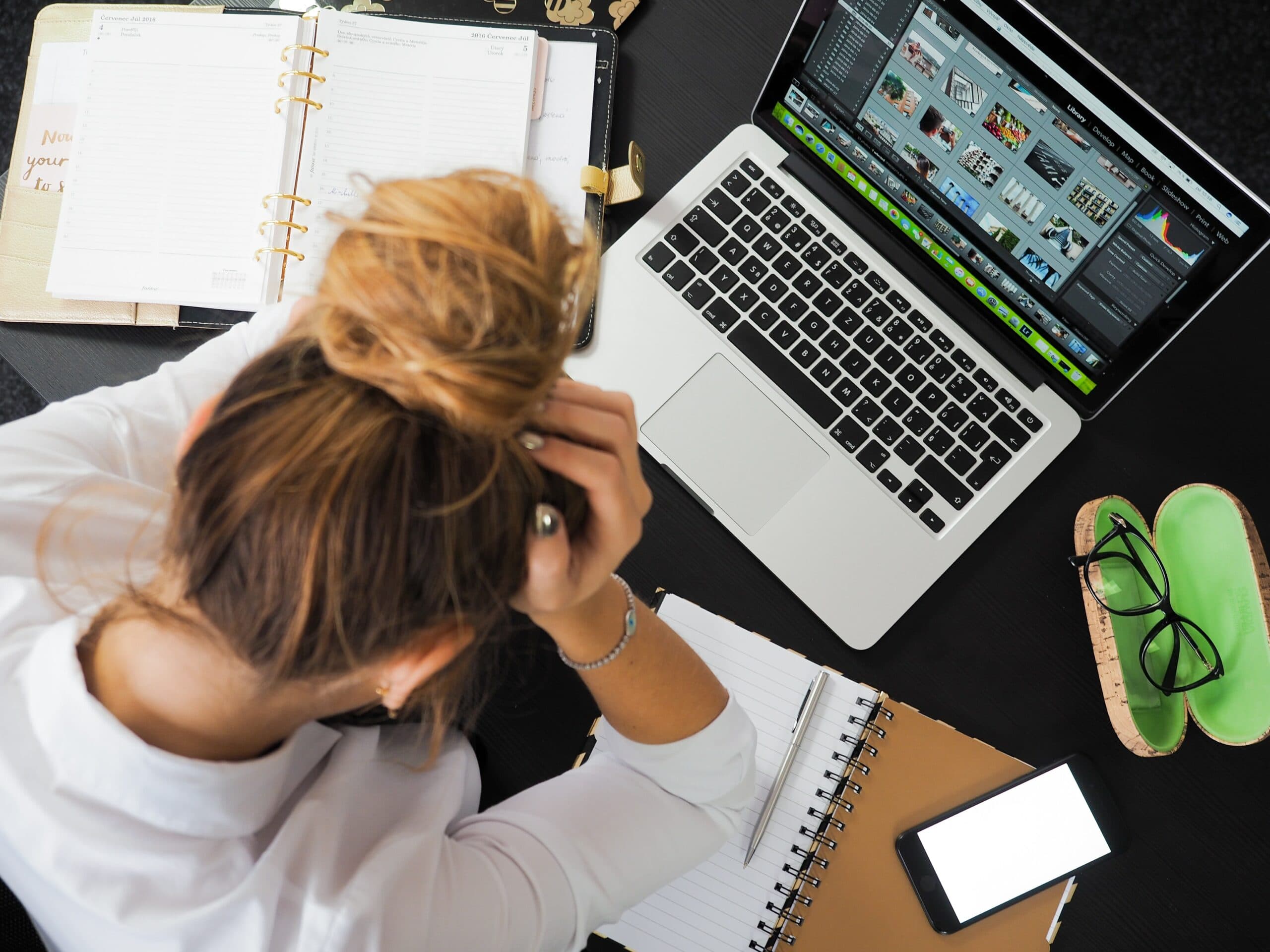 A person with tied-up hair is sitting at a desk, holding their head. An open laptop shows a photo editing program. Nearby are notebooks, a smartphone, and glasses in a green case. The scene suggests working or studying.