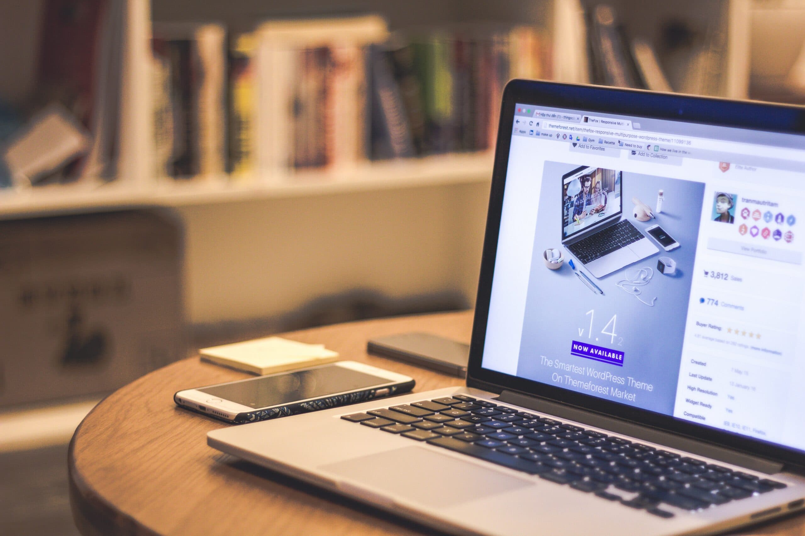 A laptop displaying a website sits on a wooden table, with a smartphone and some papers beside it. In the background, bookshelves are partially visible, creating a cozy workspace atmosphere.
