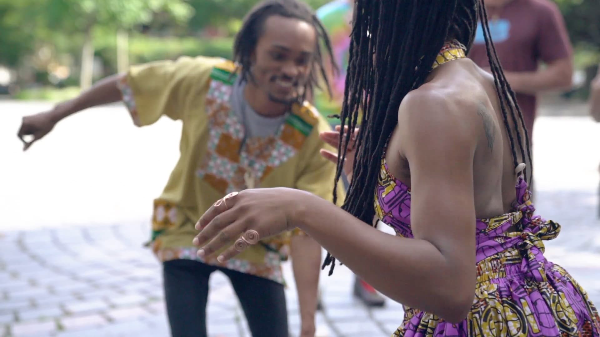 Two people dancing outdoors. A man in a yellow patterned shirt is joyfully moving, while a woman with long braids wears a purple patterned dress. They are surrounded by greenery and sunlight on cobblestone pavement.
