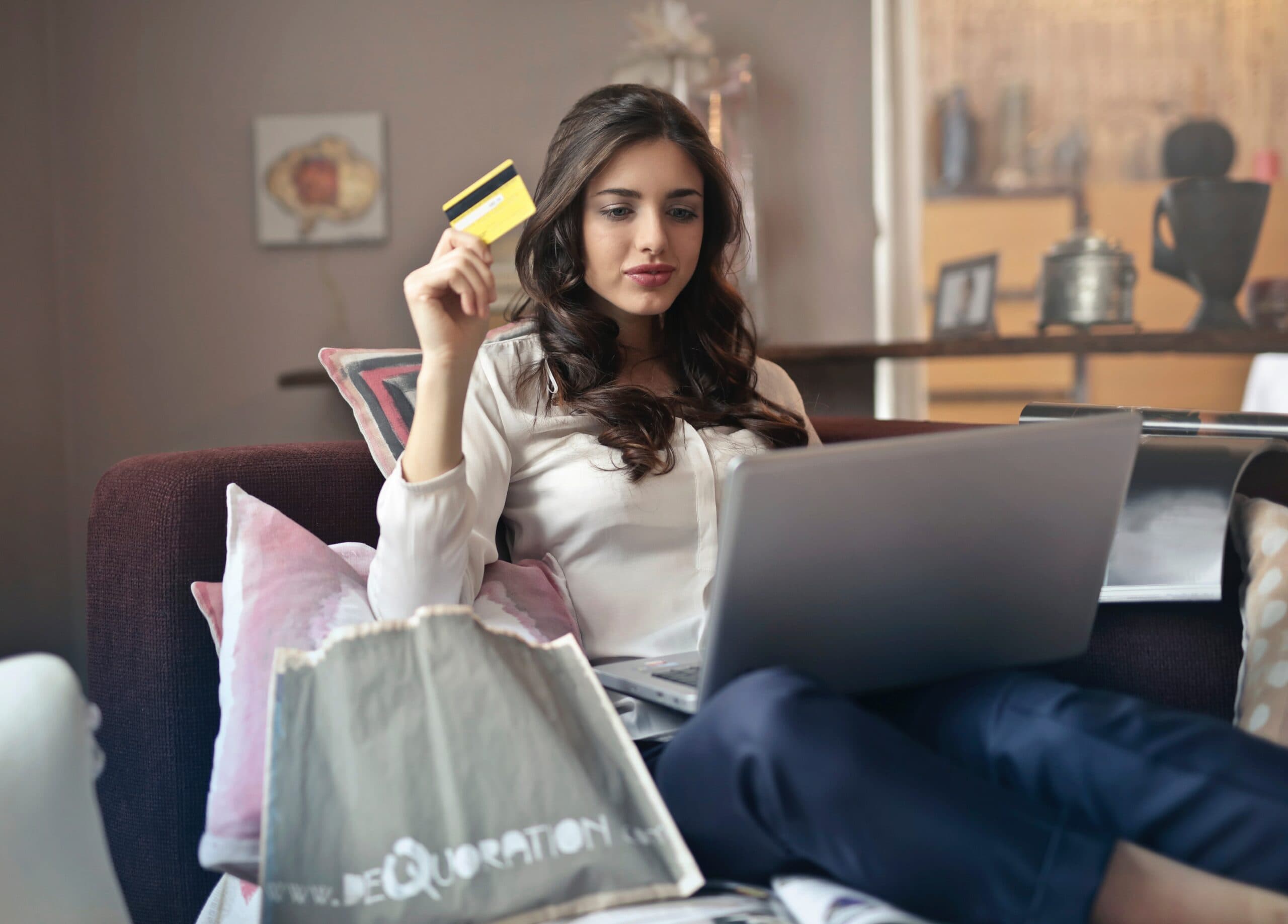 A woman with long brown hair sits on a sofa, holding a credit card in one hand and using a laptop on her lap. She is surrounded by pillows, and a shopping bag is nearby. The room has a cozy atmosphere with soft lighting.