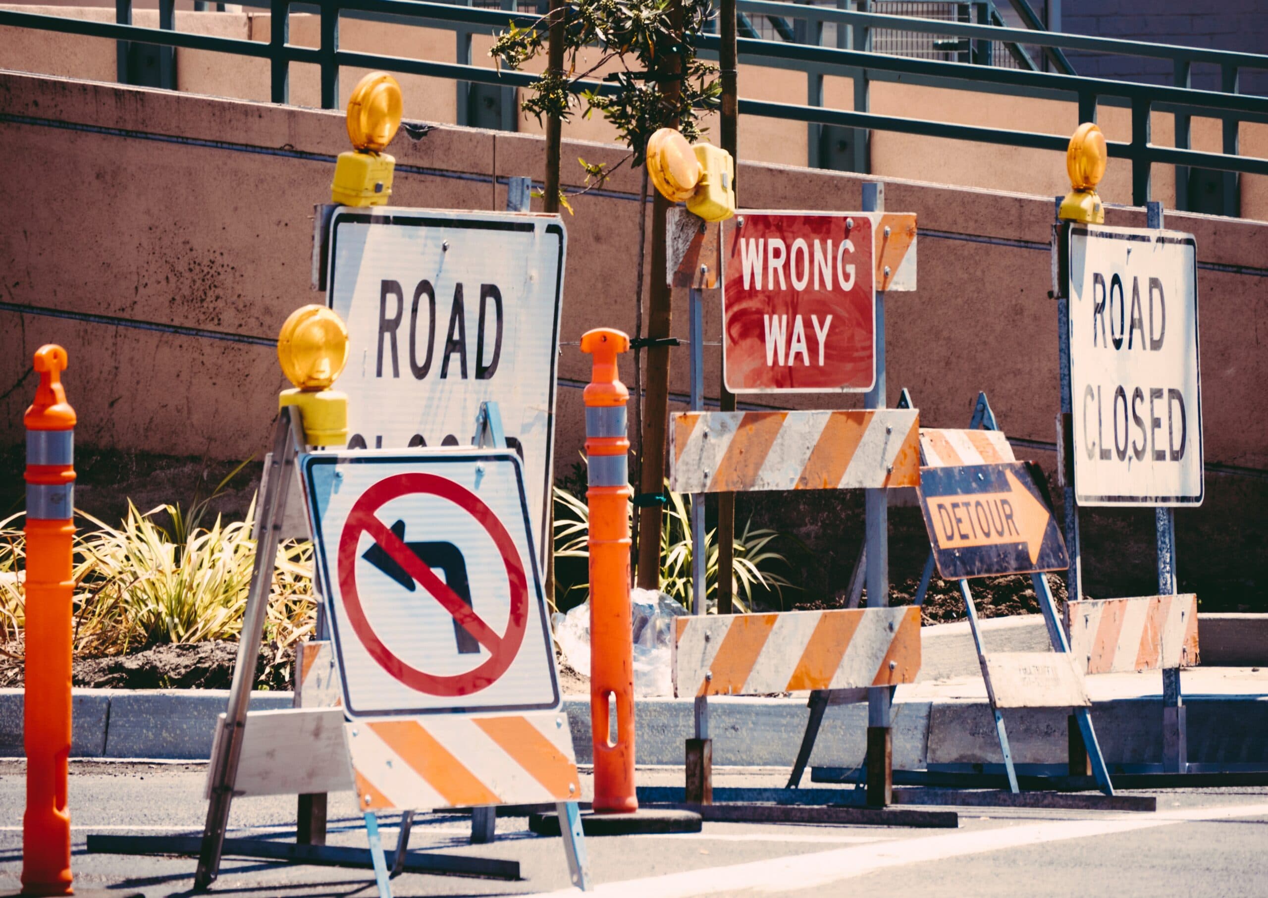 A street with multiple road signs, including Road Closed, Wrong Way, Detour, and a no left turn symbol. Orange traffic cones and barriers are also present, indicating a road closure or construction area.
