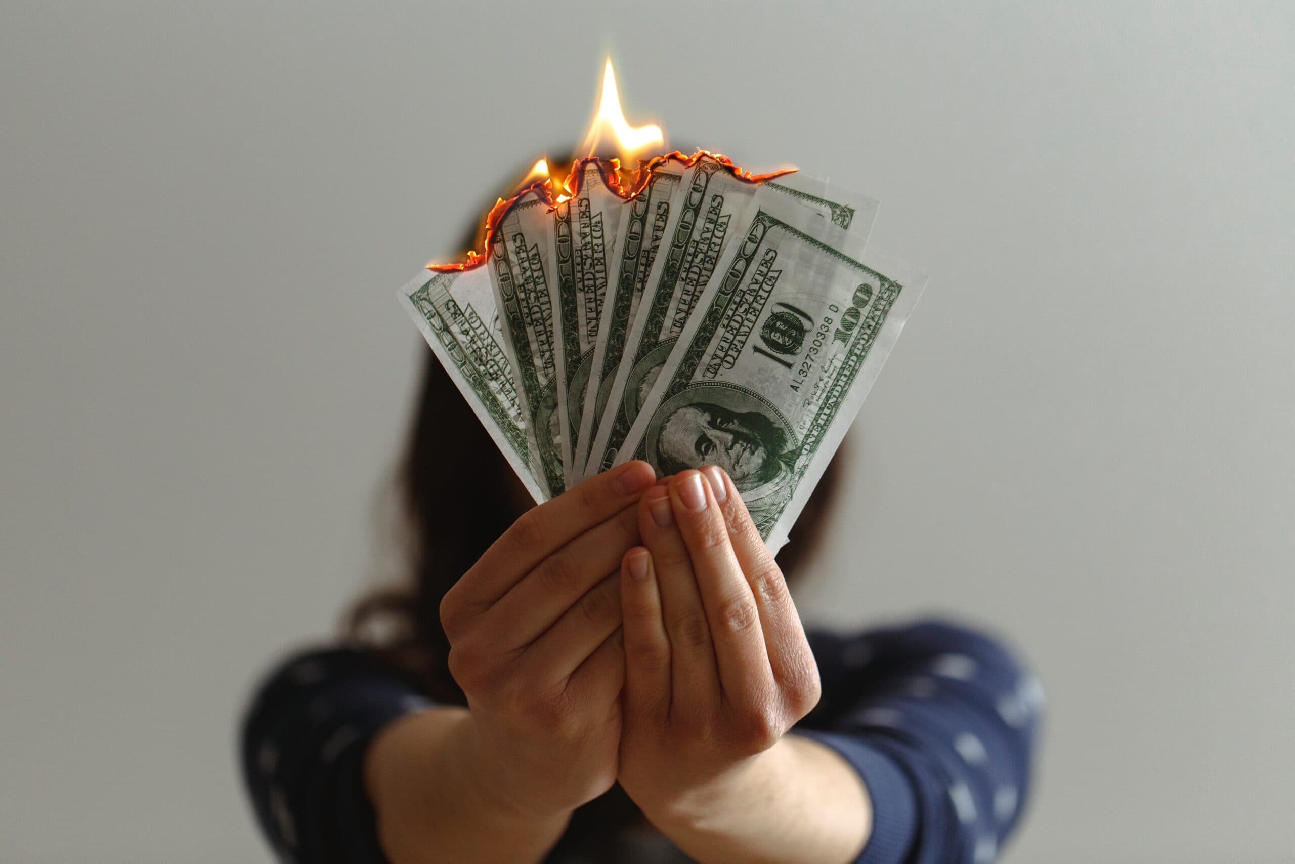 A person holding a fanned-out stack of burning U.S. hundred-dollar bills, with flames showing at the top. The individuals face is obscured, and they are wearing a dark blue long-sleeve shirt with white details against a light gray background.